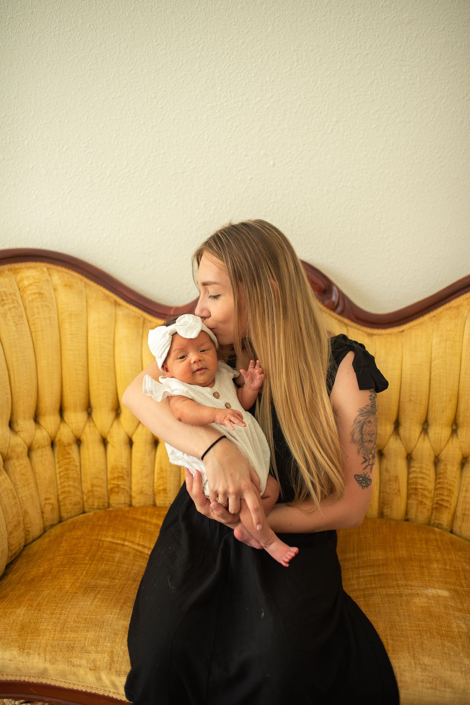 A blonde mother in a black dress kisses her newborn baby on a vintage gold sofa.