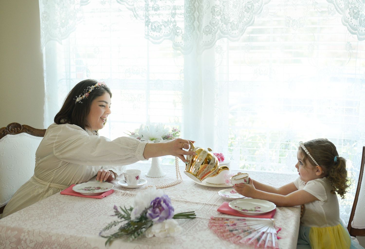 Woman in white dress passing treats to a young girl at an elegant tea party table with fine china.