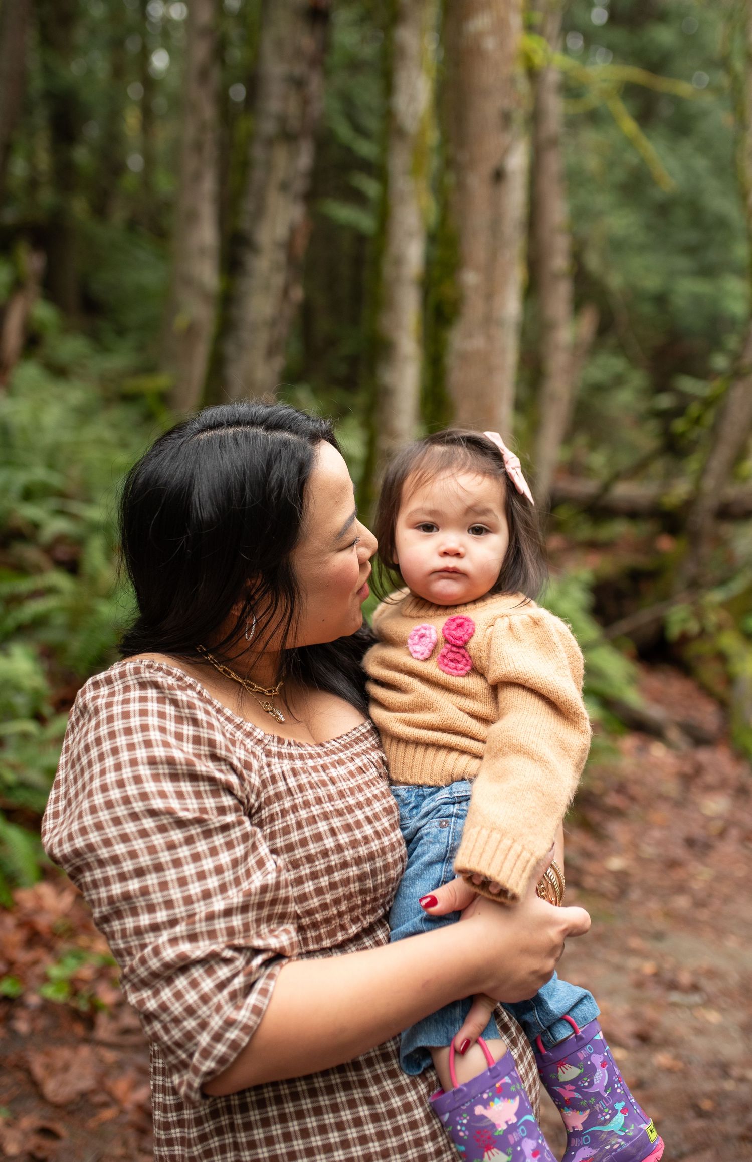 A mother lovingly holds her toddler daughter in a lush forest setting, both wearing warm autumn outfits.