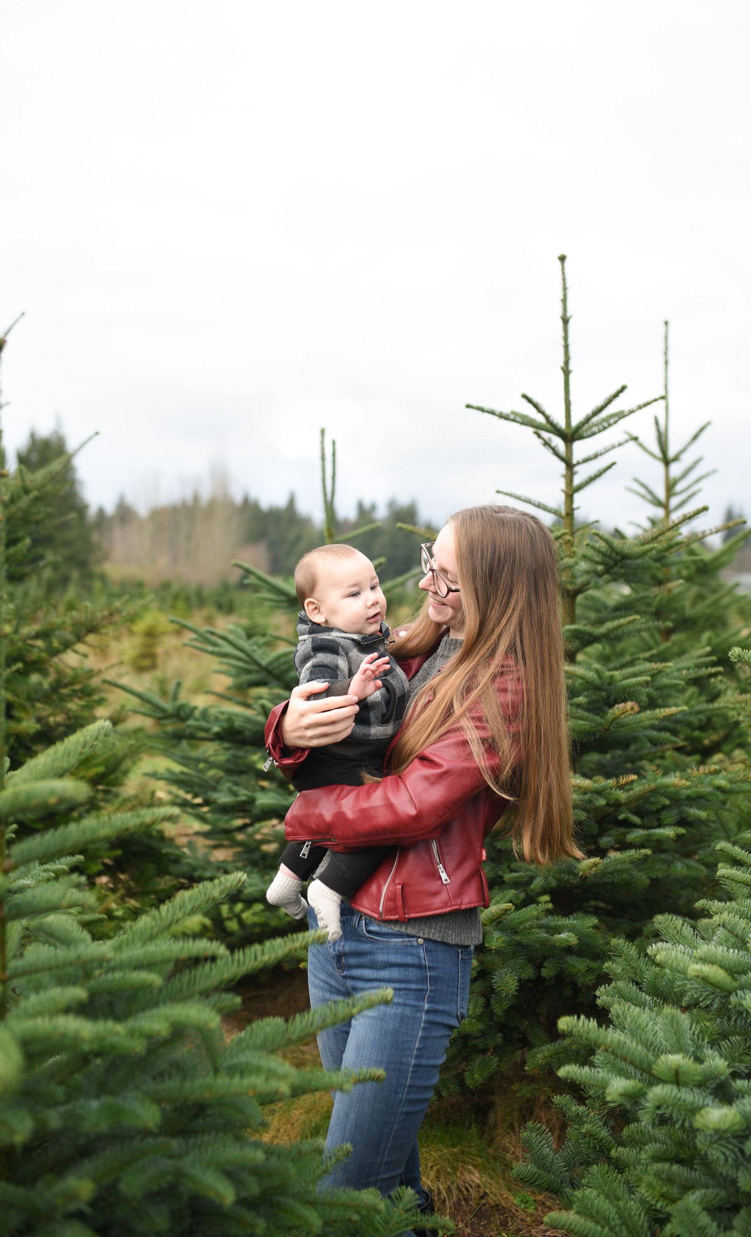 A young mother in a red jacket holds her baby close while standing among green Christmas trees.