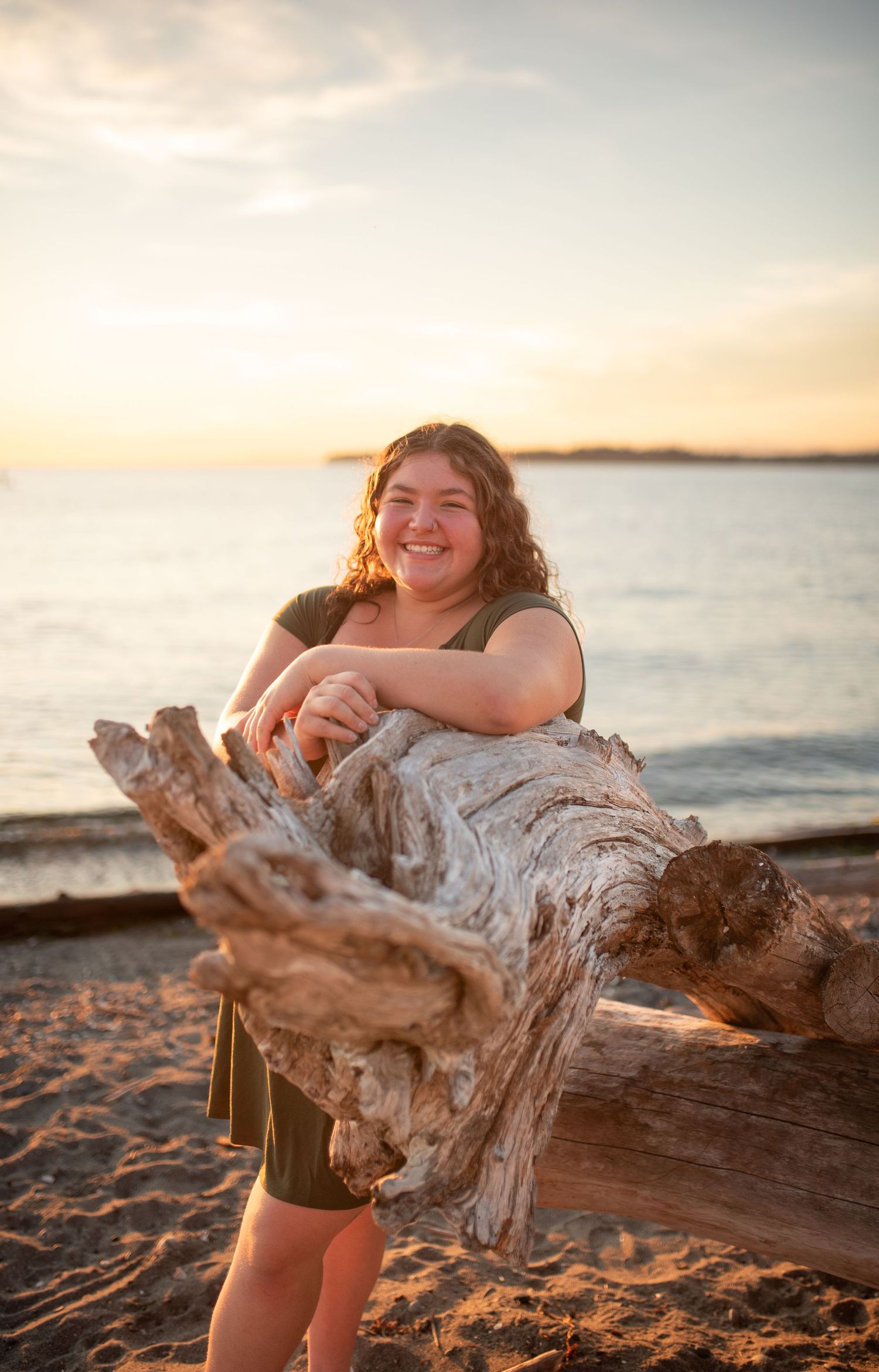 Young woman smiling and leaning on driftwood log at sandy beach during golden sunset.