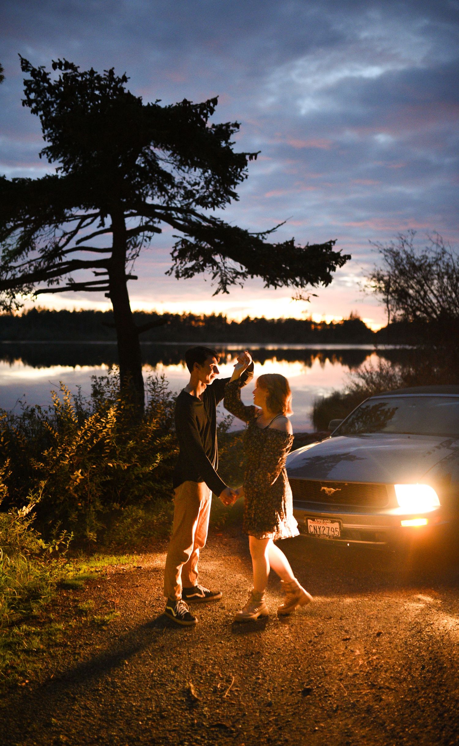 Couple dancing beside a Mustang car at dusk near a serene lake with dramatic cloudy sky.