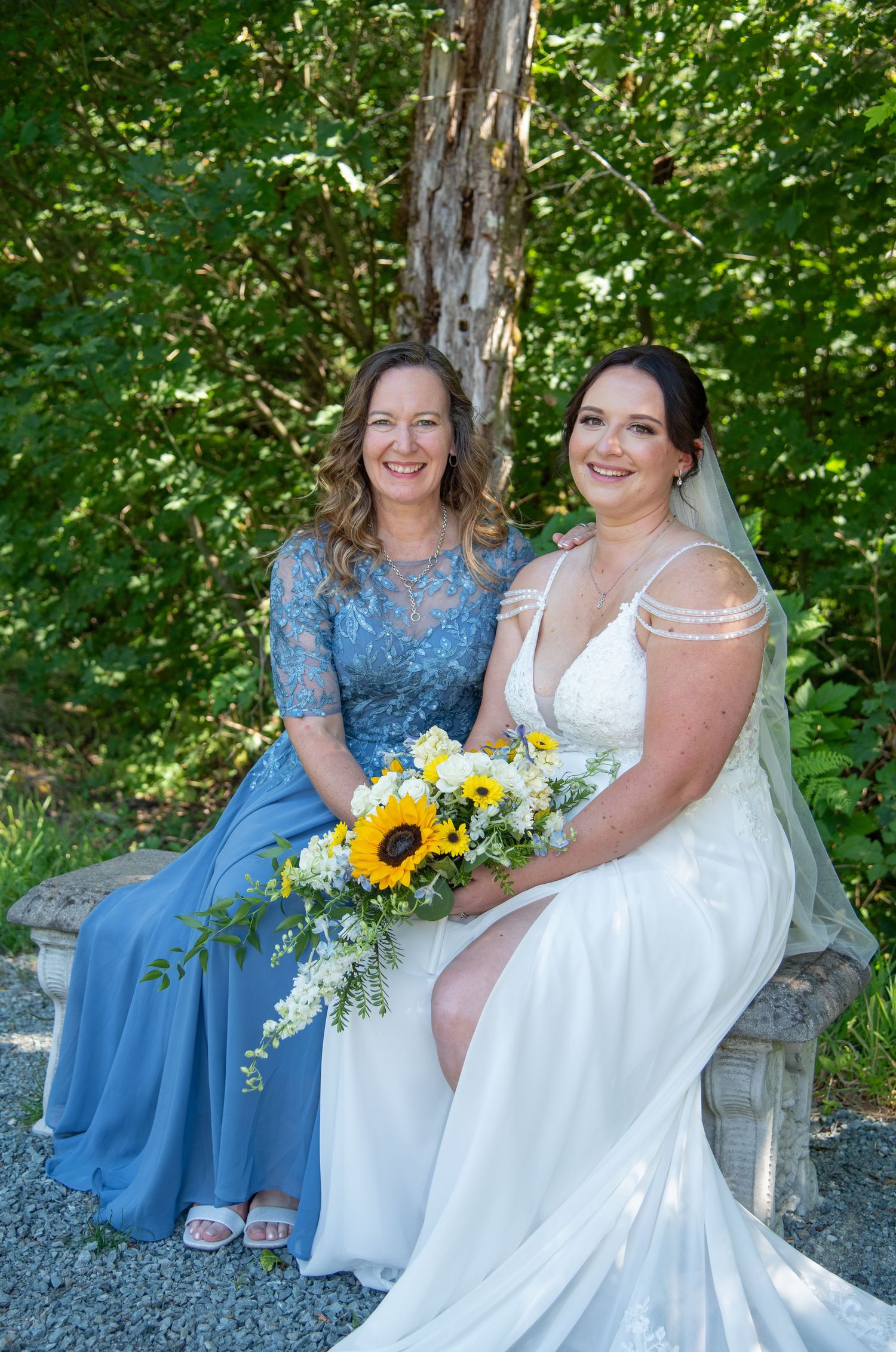 A bride in a white gown holds sunflowers while posing with a woman in a blue lace dress.