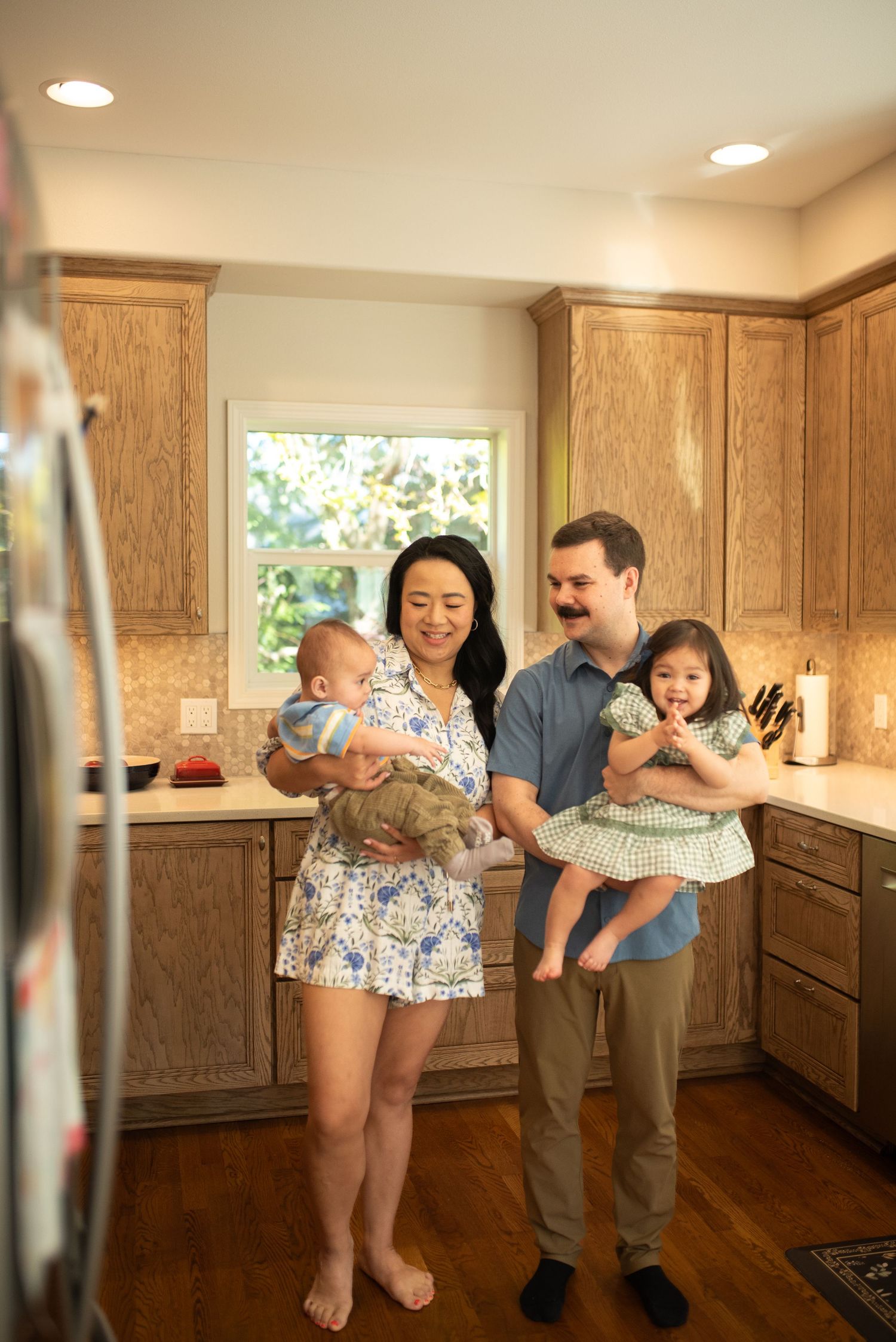 Happy family of four posing in a warm kitchen, with parents holding a baby and a toddler girl.