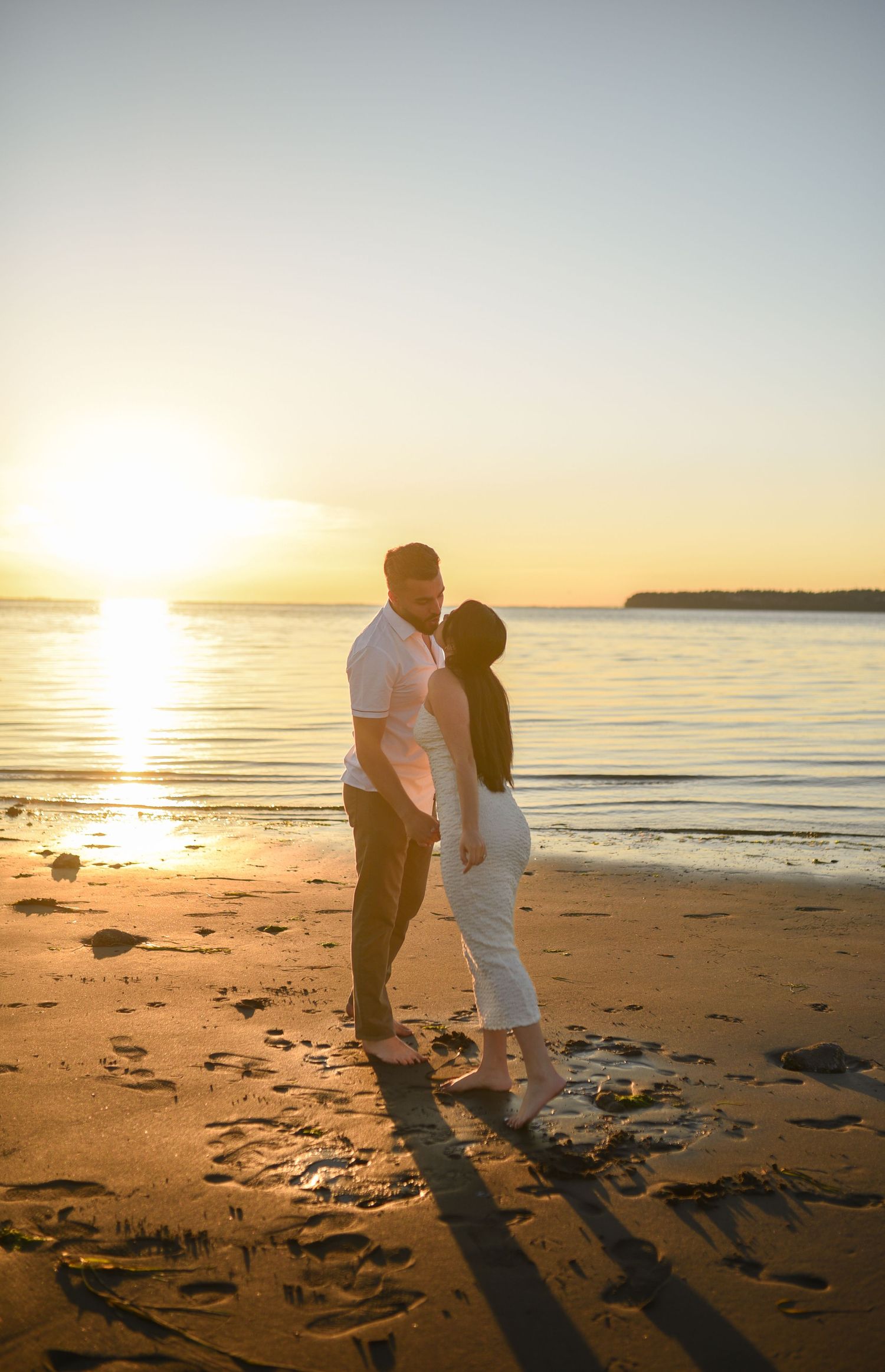 Couple sharing a romantic moment on a sandy beach at golden sunset with calm ocean waters in the background.