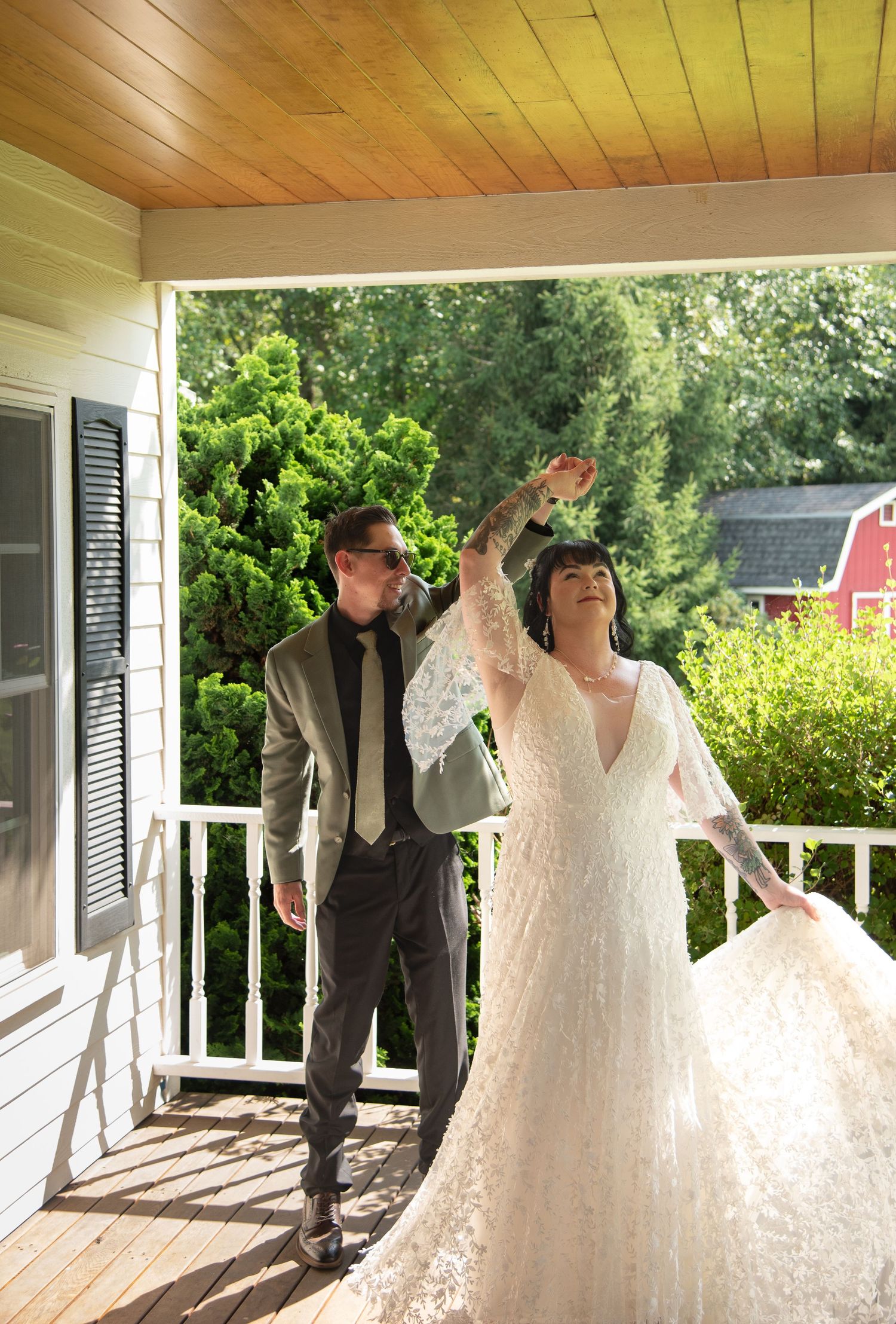 Groom twirls bride in lace wedding gown on sunlit porch with green trees in background.