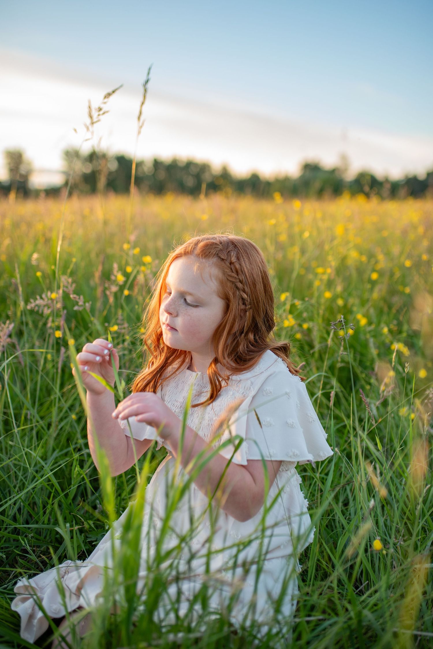 Young red-haired girl in white dress sits in a wildflower meadow examining tall grass at sunset.