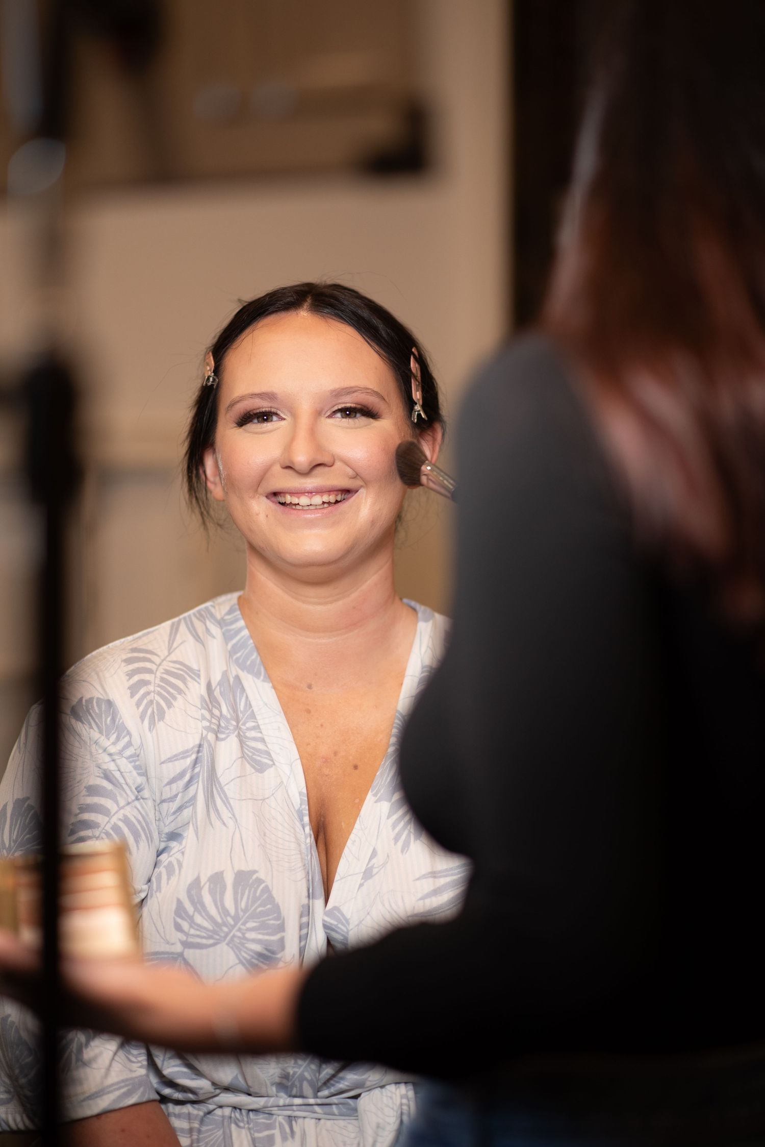A smiling woman in a floral wrap dress gets her makeup done by a professional artist.