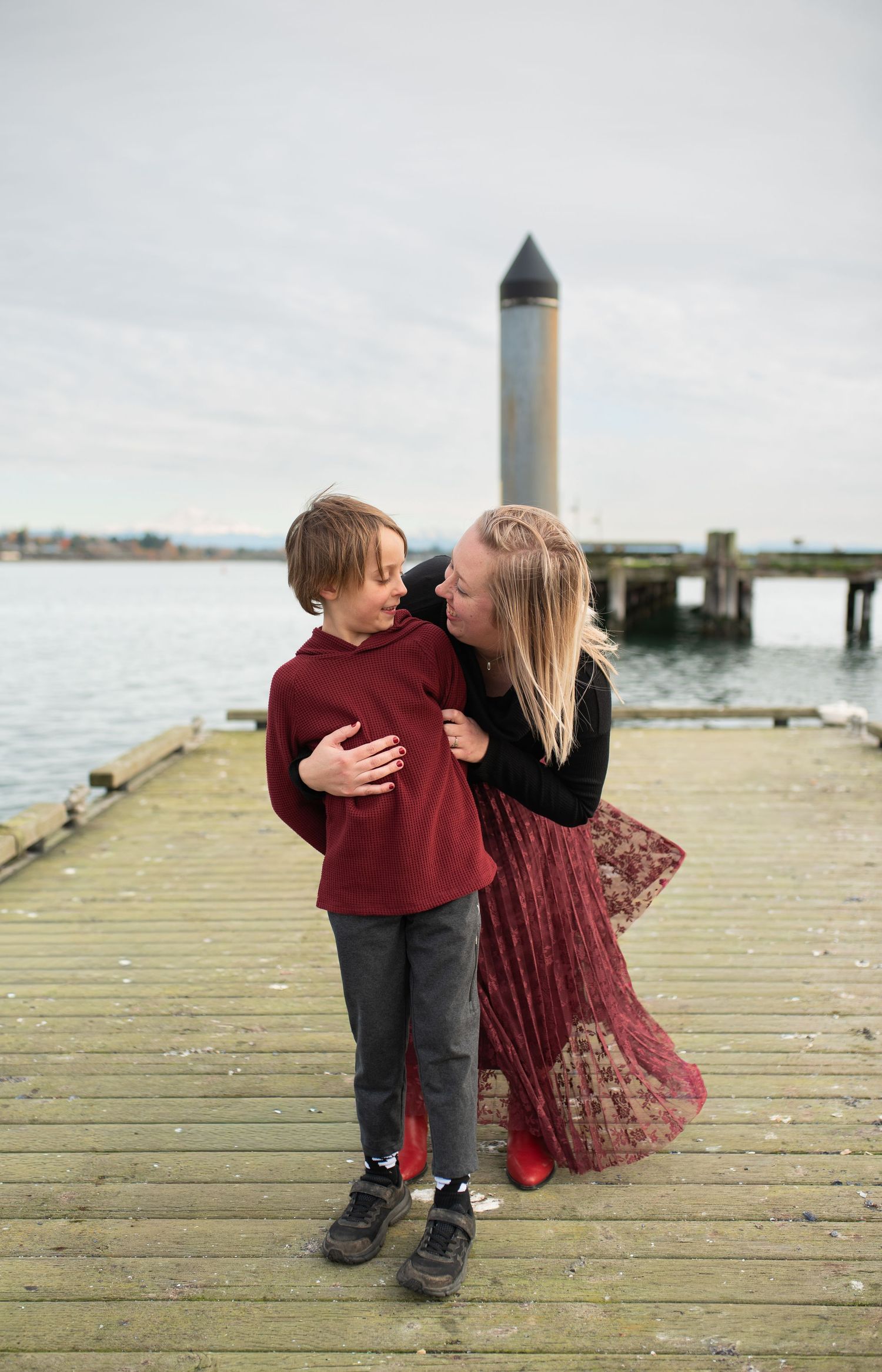 A mother and young boy share a warm embrace on a wooden pier overlooking the water.