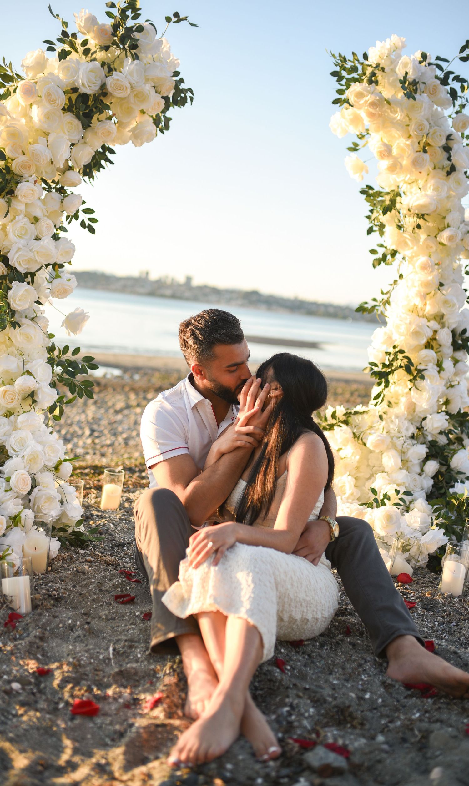 Couple sharing a romantic moment on a beach under a white floral arch with candles at sunset.
