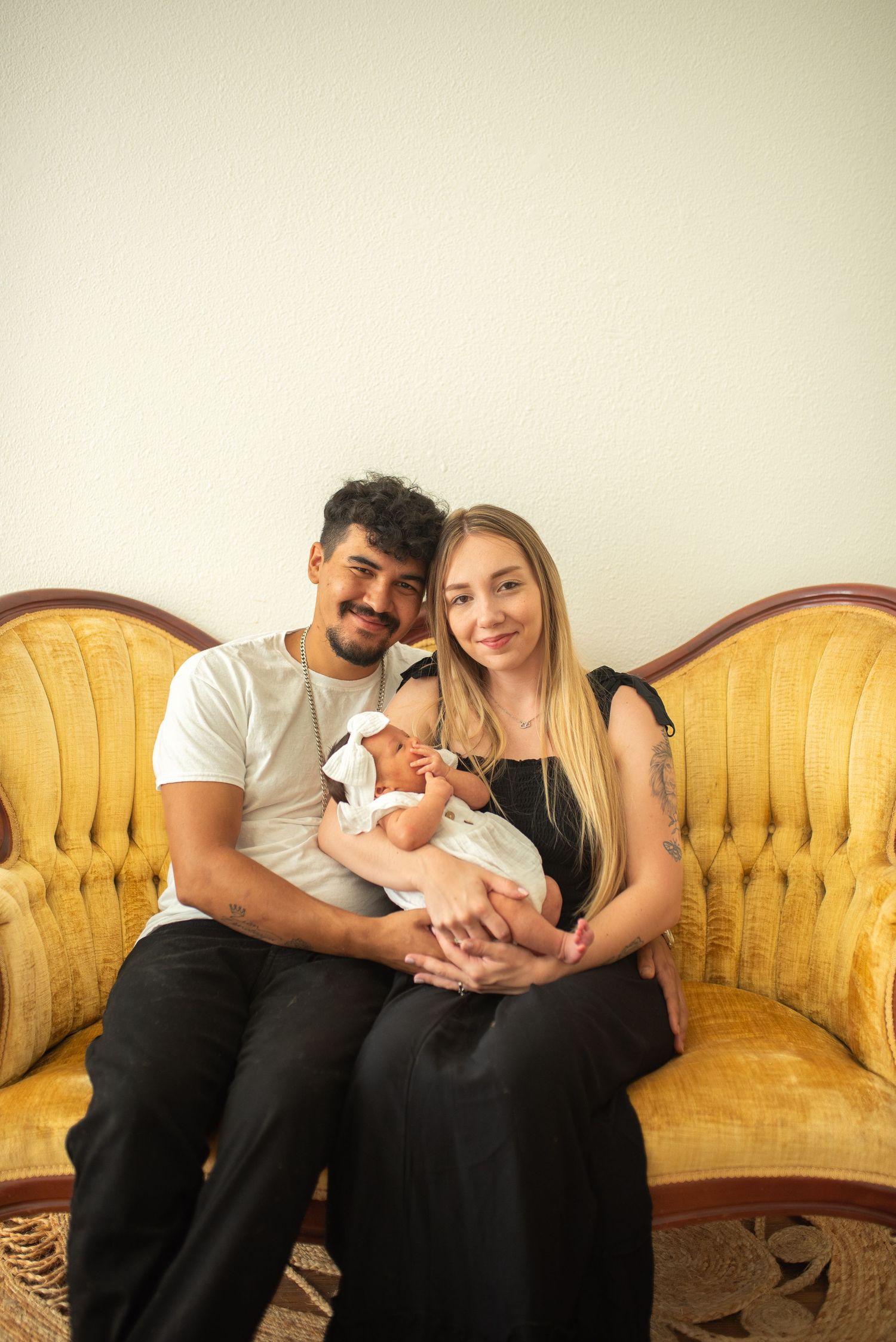 Smiling young couple sitting on vintage gold sofa proudly holding their newborn baby in white.