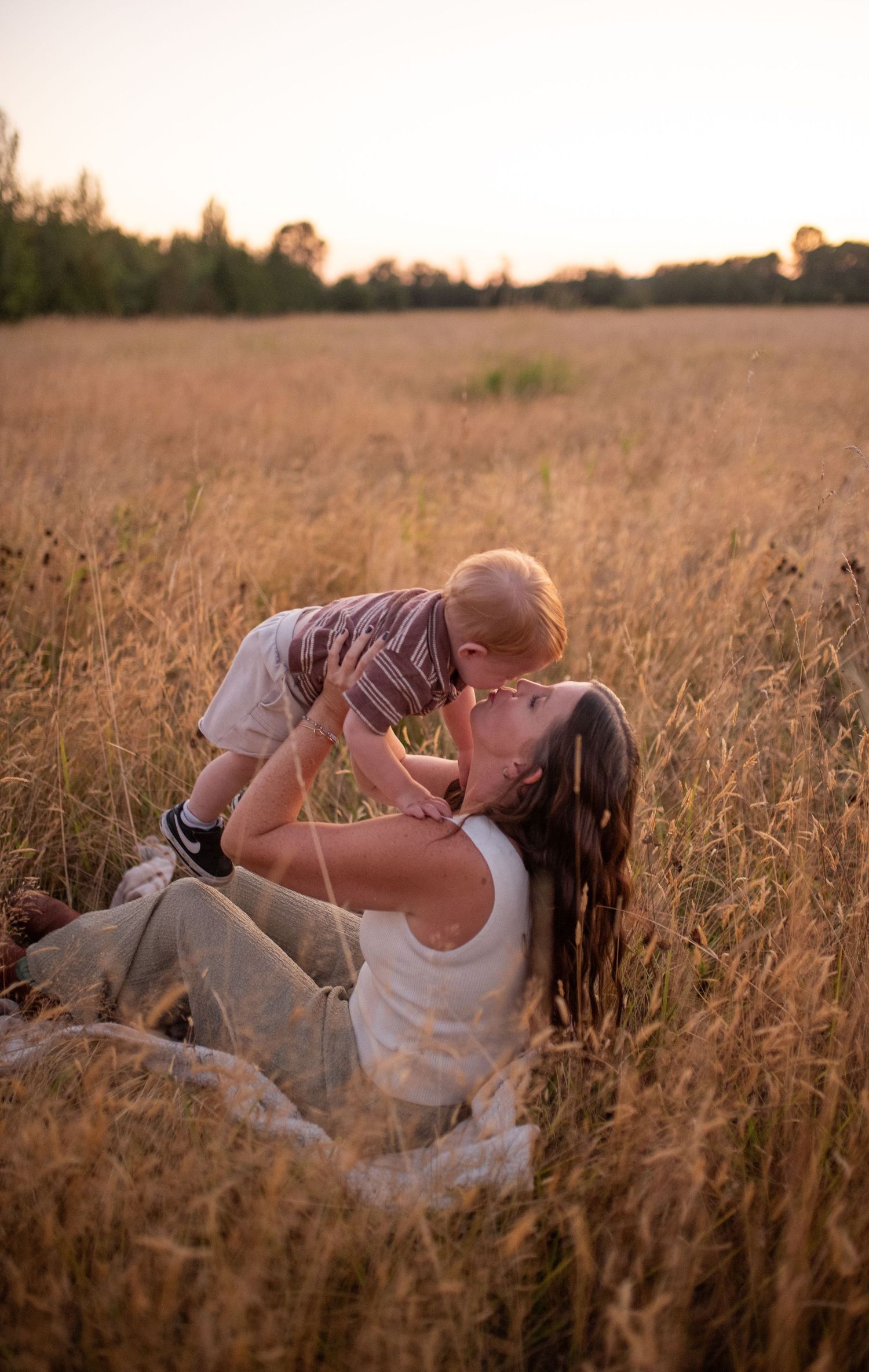 Mother sitting in golden field lifting baby for a kiss during warm sunset light.
