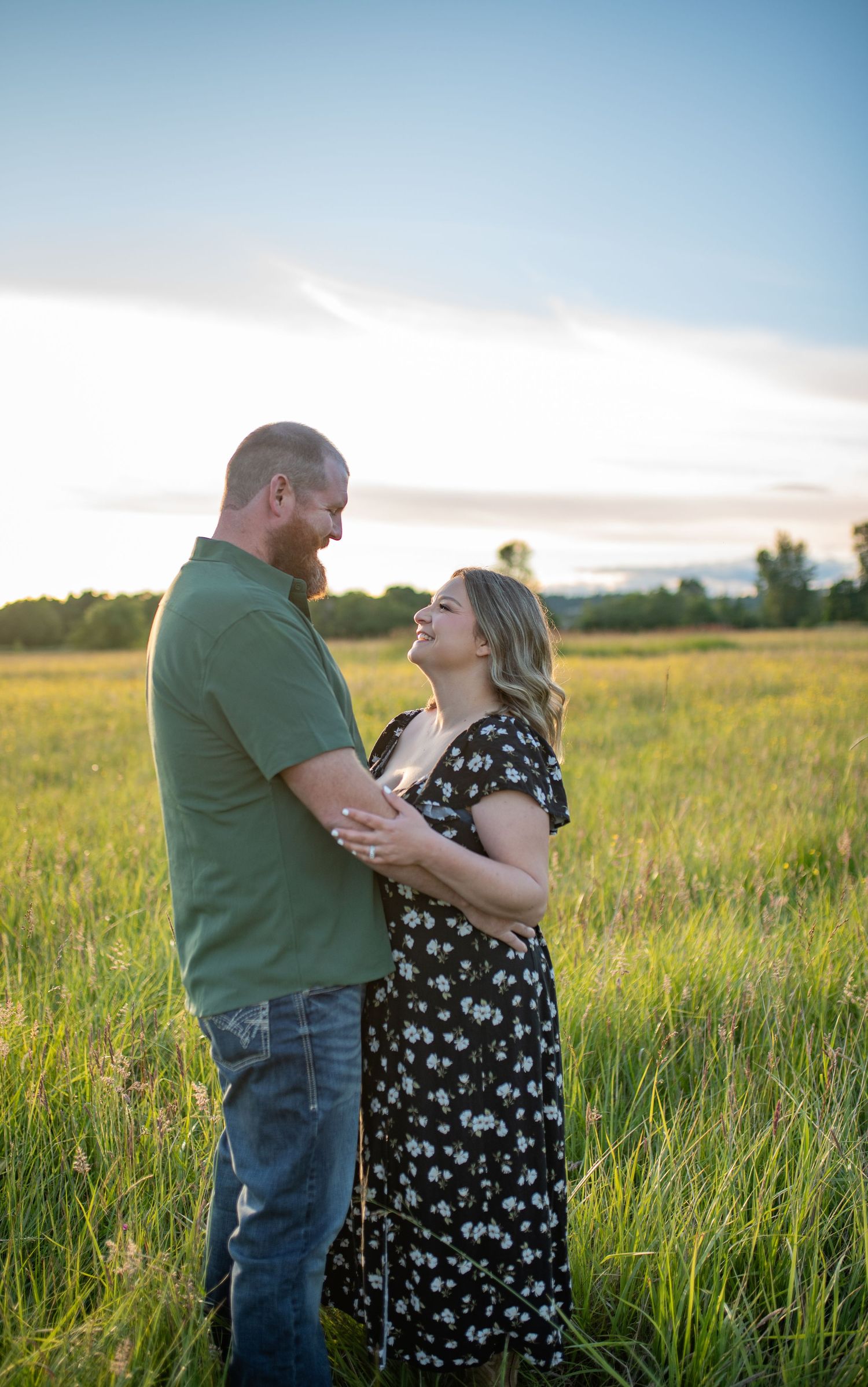 Couple embracing and smiling at each other in a sunny green field at golden hour, man in green shirt and woman in floral dress.