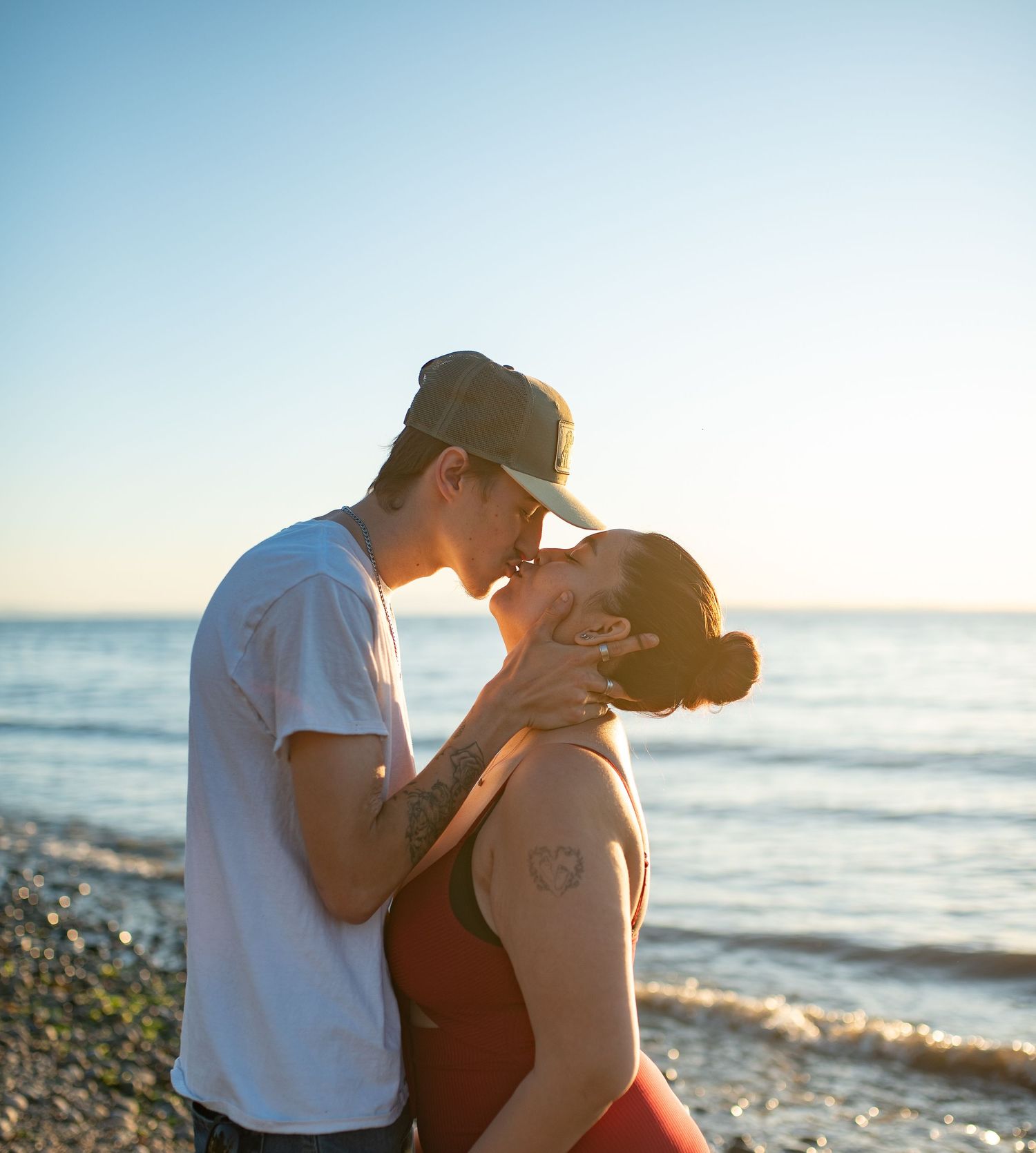 Couple sharing a kiss on a pebble beach at sunset, man in cap and woman in red dress.