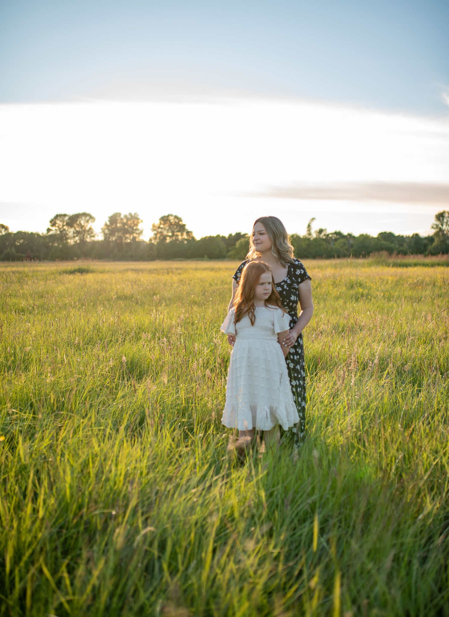 Mother in floral dress embraces red-haired daughter in white dress standing in a sunlit green meadow.
