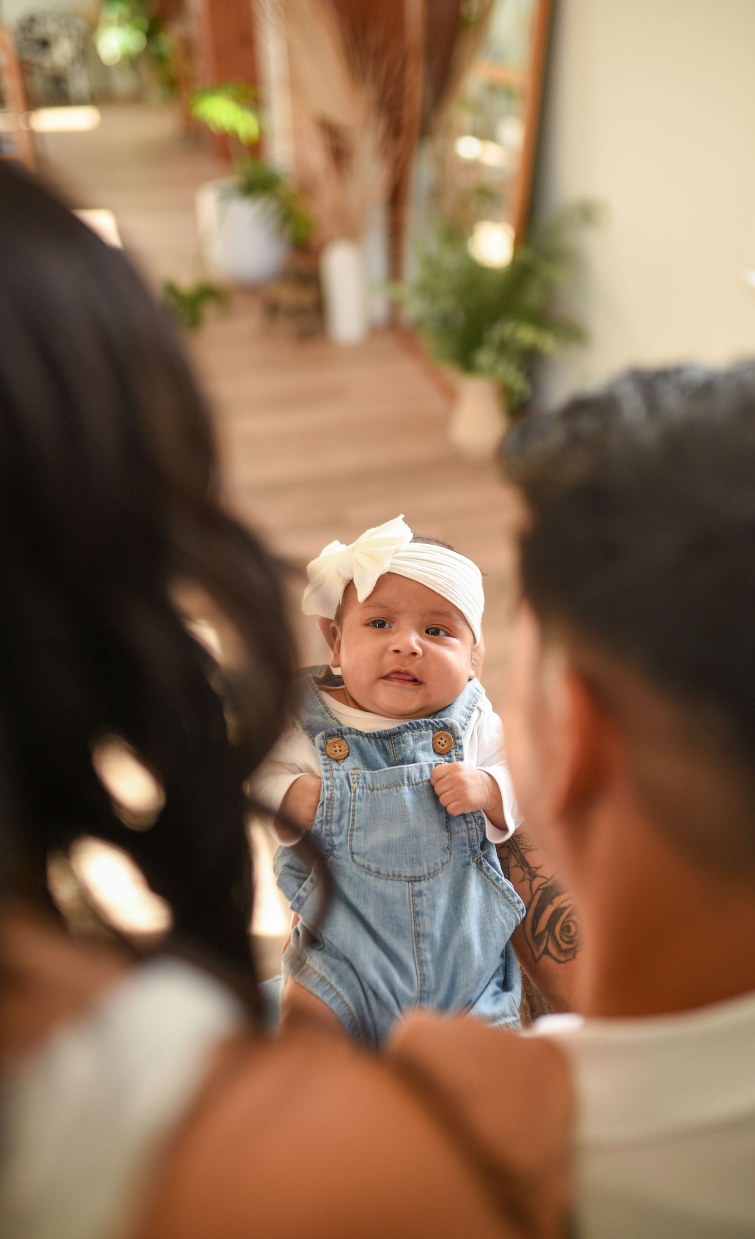 Parents holding their newborn baby girl in denim overalls and white bow headband in a bright indoor setting.