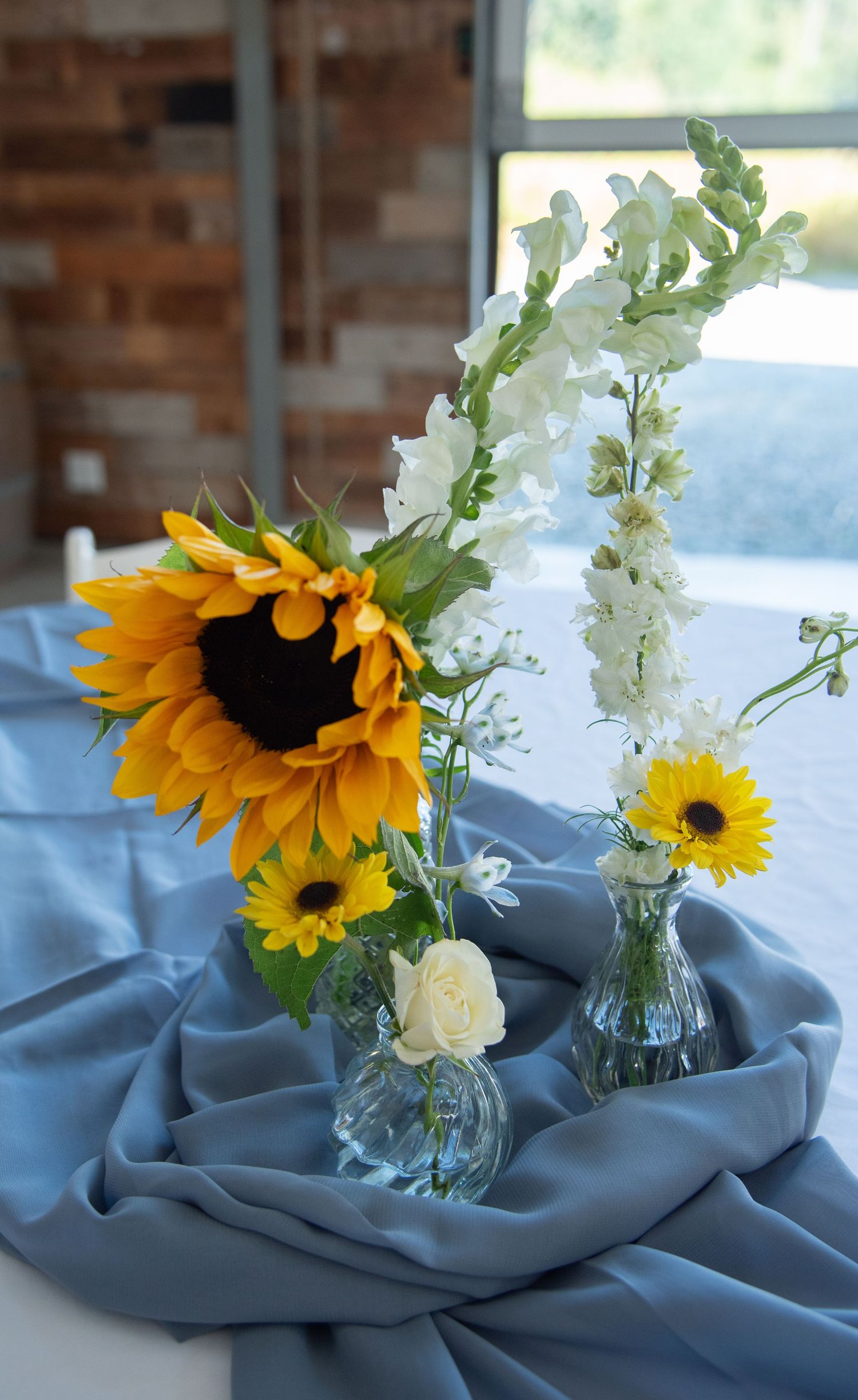 Elegant wedding table centerpiece featuring sunflowers and white blooms in glass vases on a dusty blue draped table.