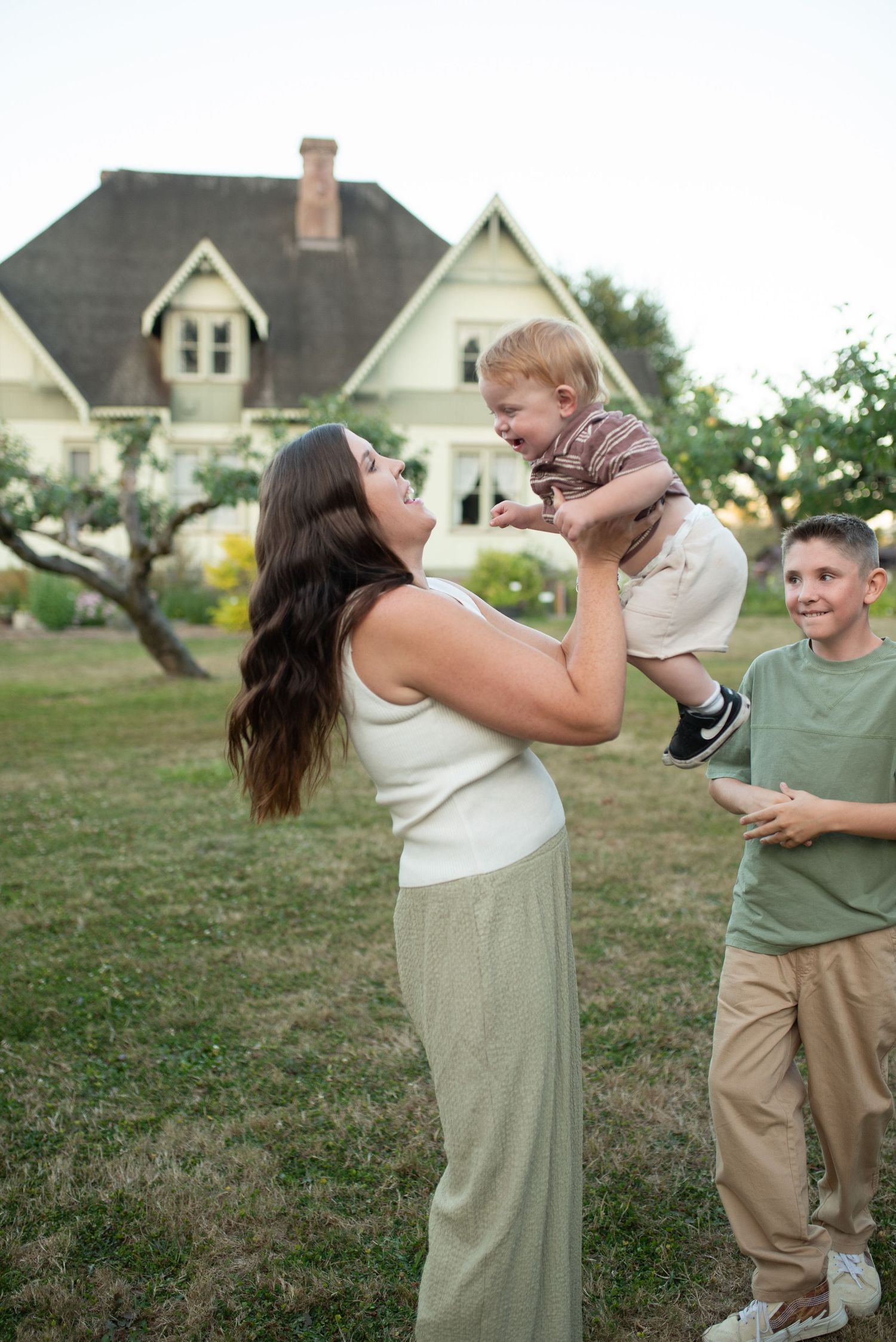A mother lifts her laughing baby in the air while an older boy watches in a garden near a Victorian home.
