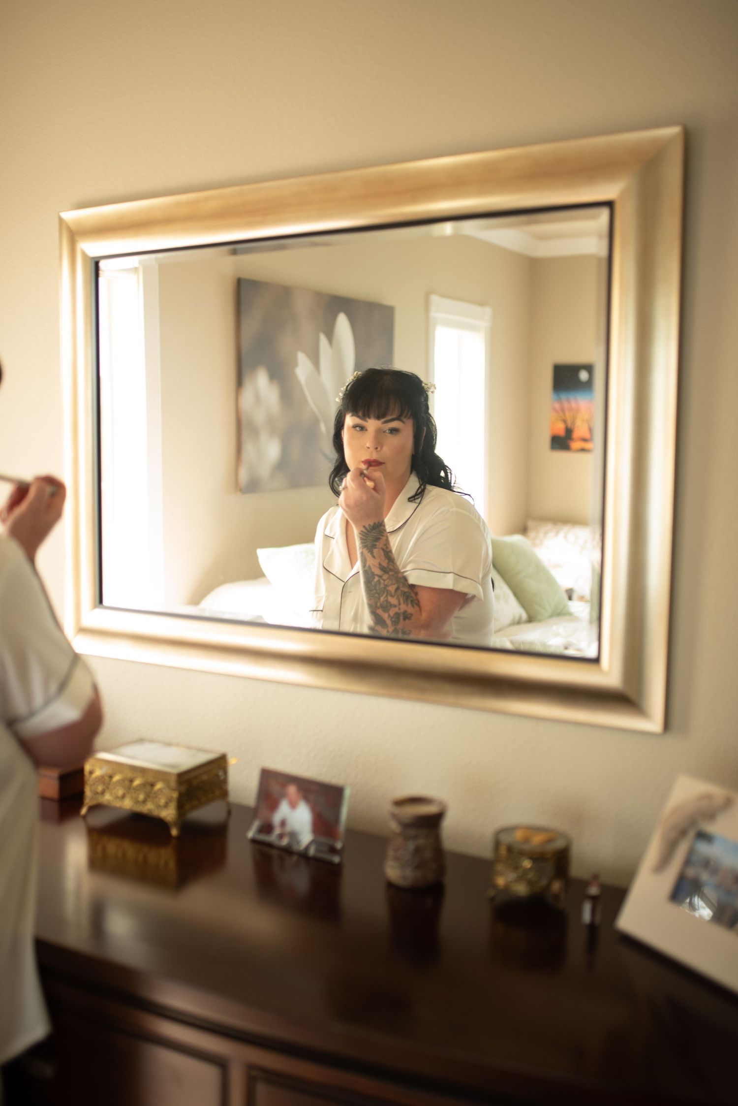 A tattooed woman applies makeup while gazing into a gold-framed mirror atop a wooden dresser in a bedroom.