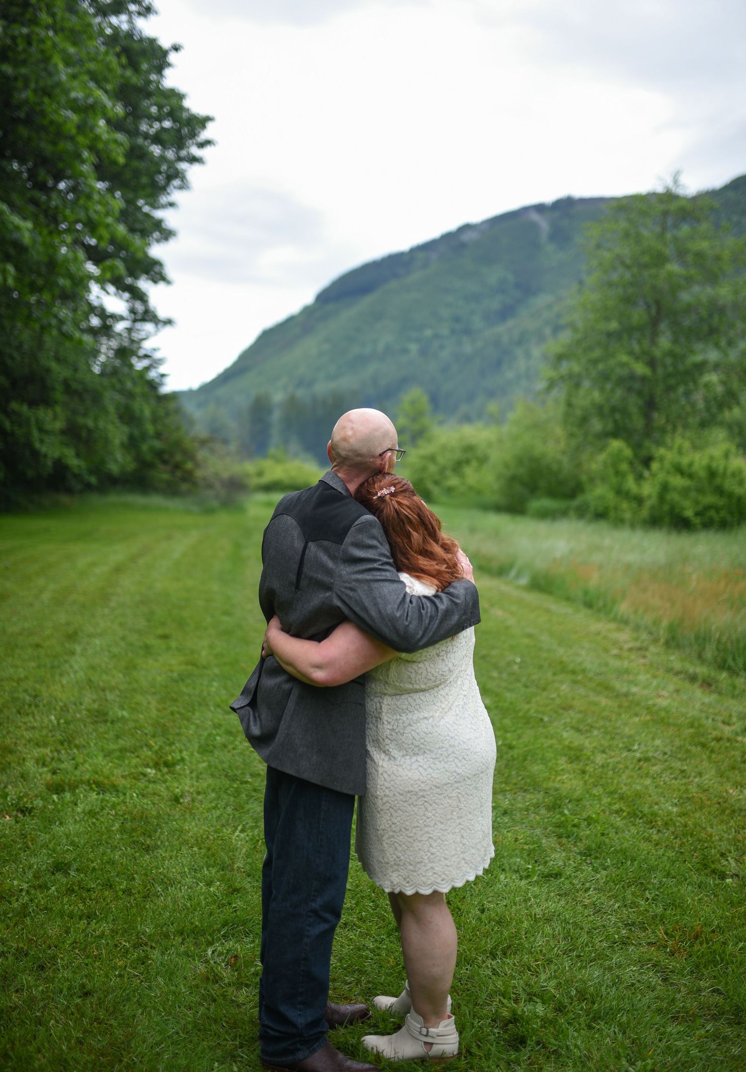 A couple embraces in a lush green meadow with mountain views, the woman in a white lace dress and the man in a gray suit.