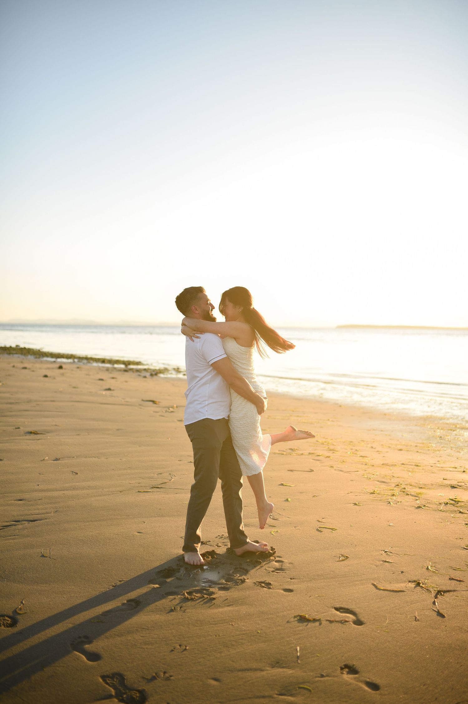 Couple embracing on a sunny beach at golden hour, man lifting woman in white dress with ocean in the background.