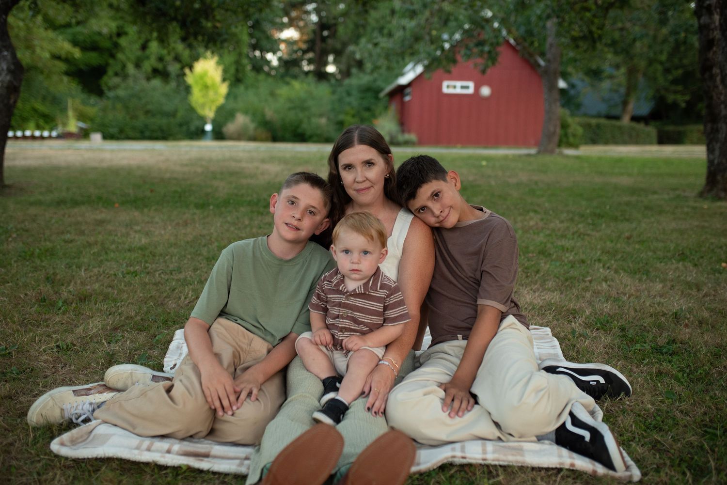 A mother sits on a blanket in a park with her three sons, smiling warmly for a family portrait.
