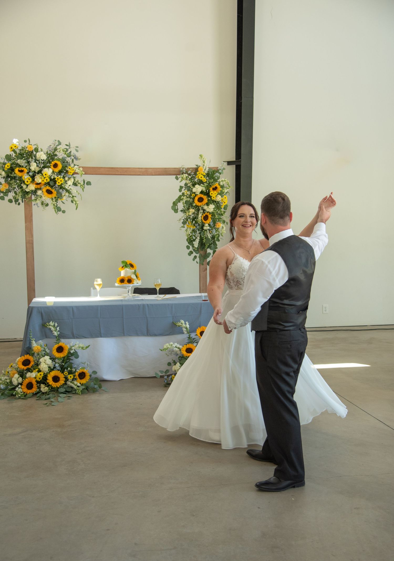 Bride and groom sharing their first dance in a decorated venue with a sunflower arch and blue-draped sweetheart table.