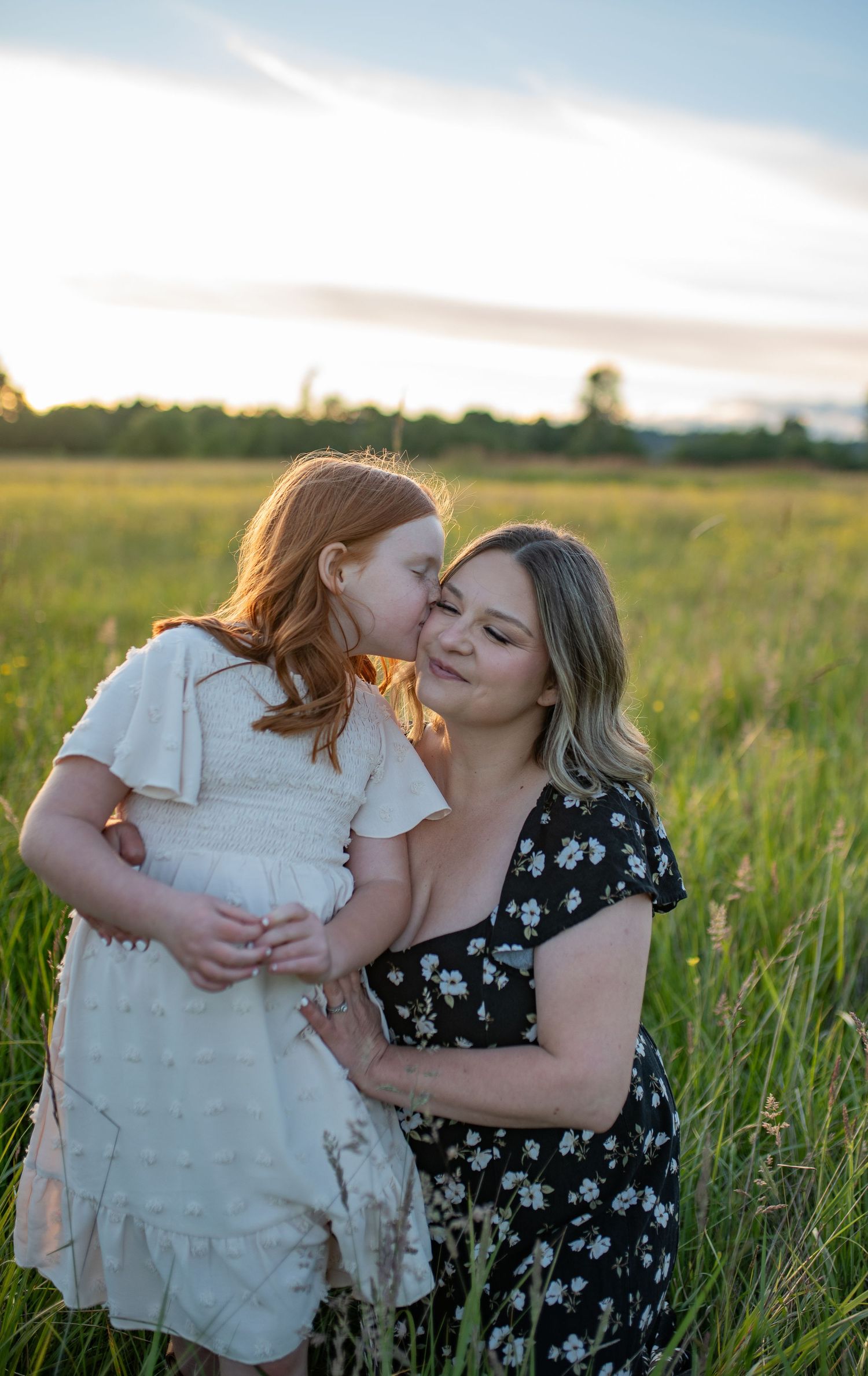 Red-haired girl kisses mother on cheek while being held in a golden-lit grassy field at sunset.