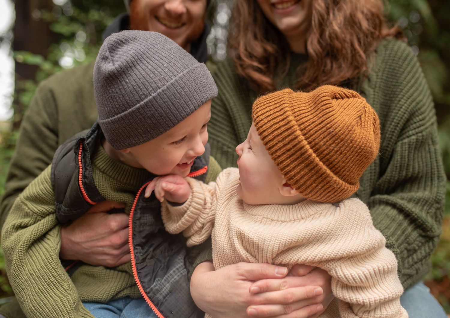 Two young siblings in knit beanies smile at each other while being held by their parents.