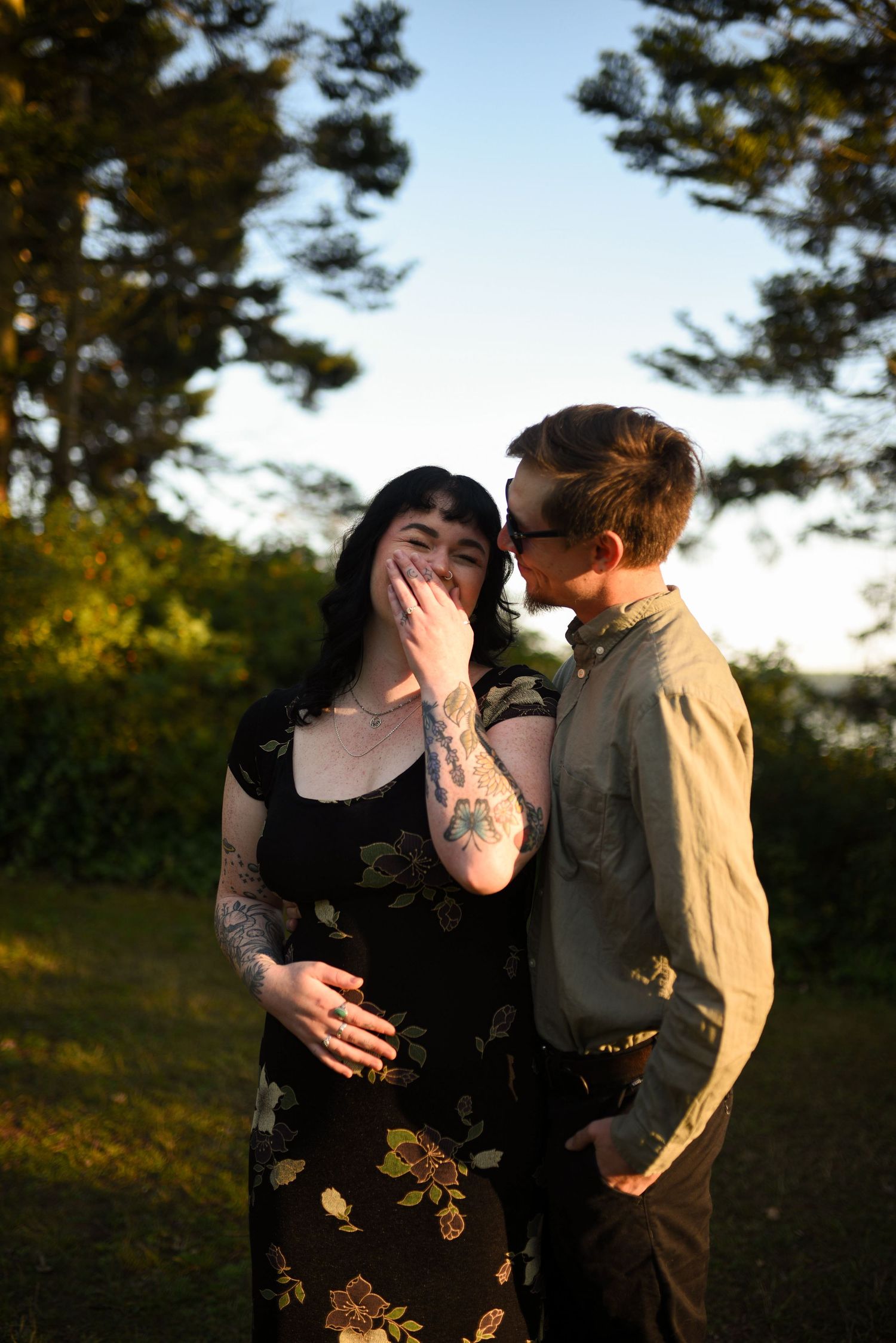Woman laughing joyfully with tattooed arm while man whispers to her in sunny park.