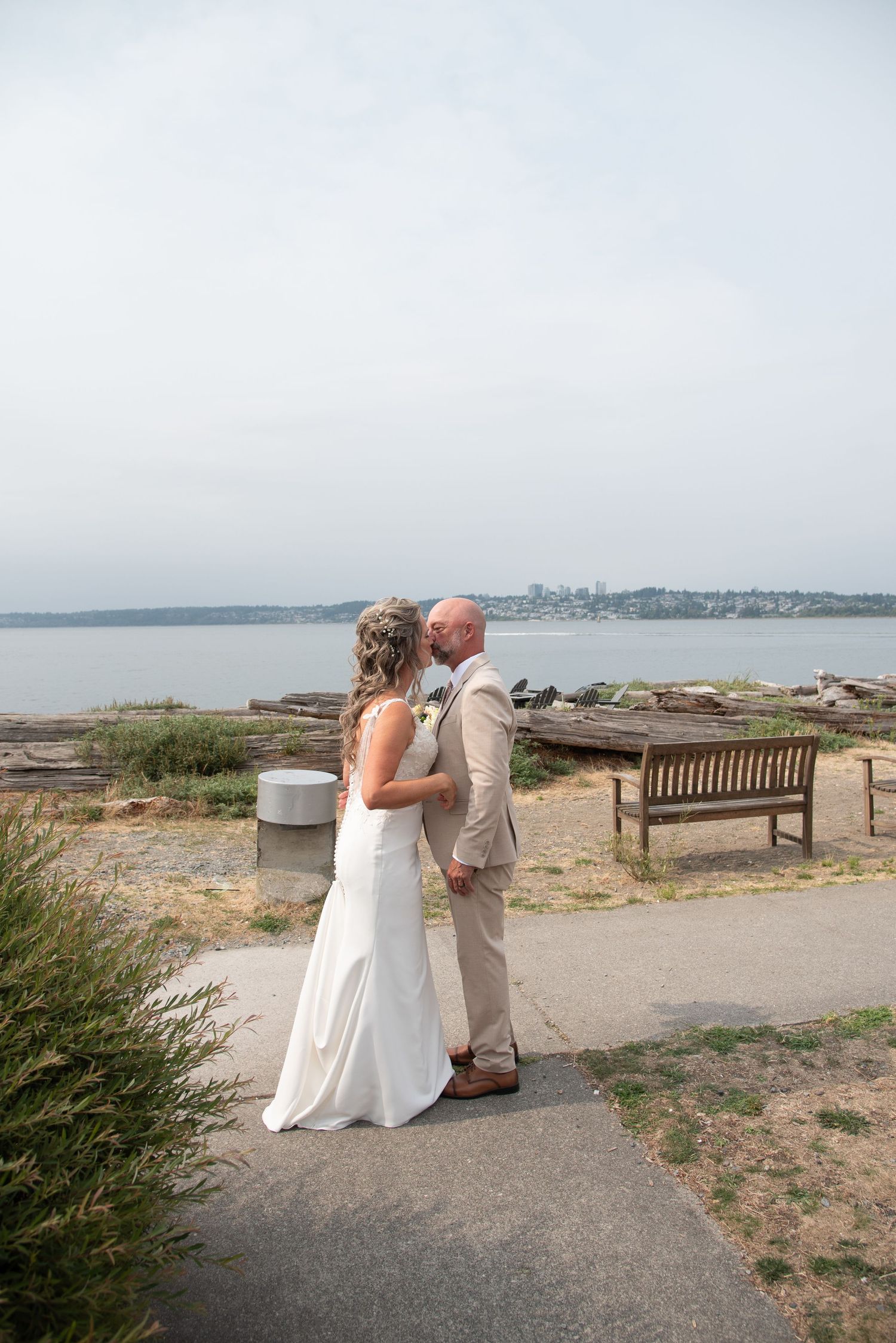 Bride and groom share a romantic kiss during first look on scenic waterfront path with bench and ocean view.