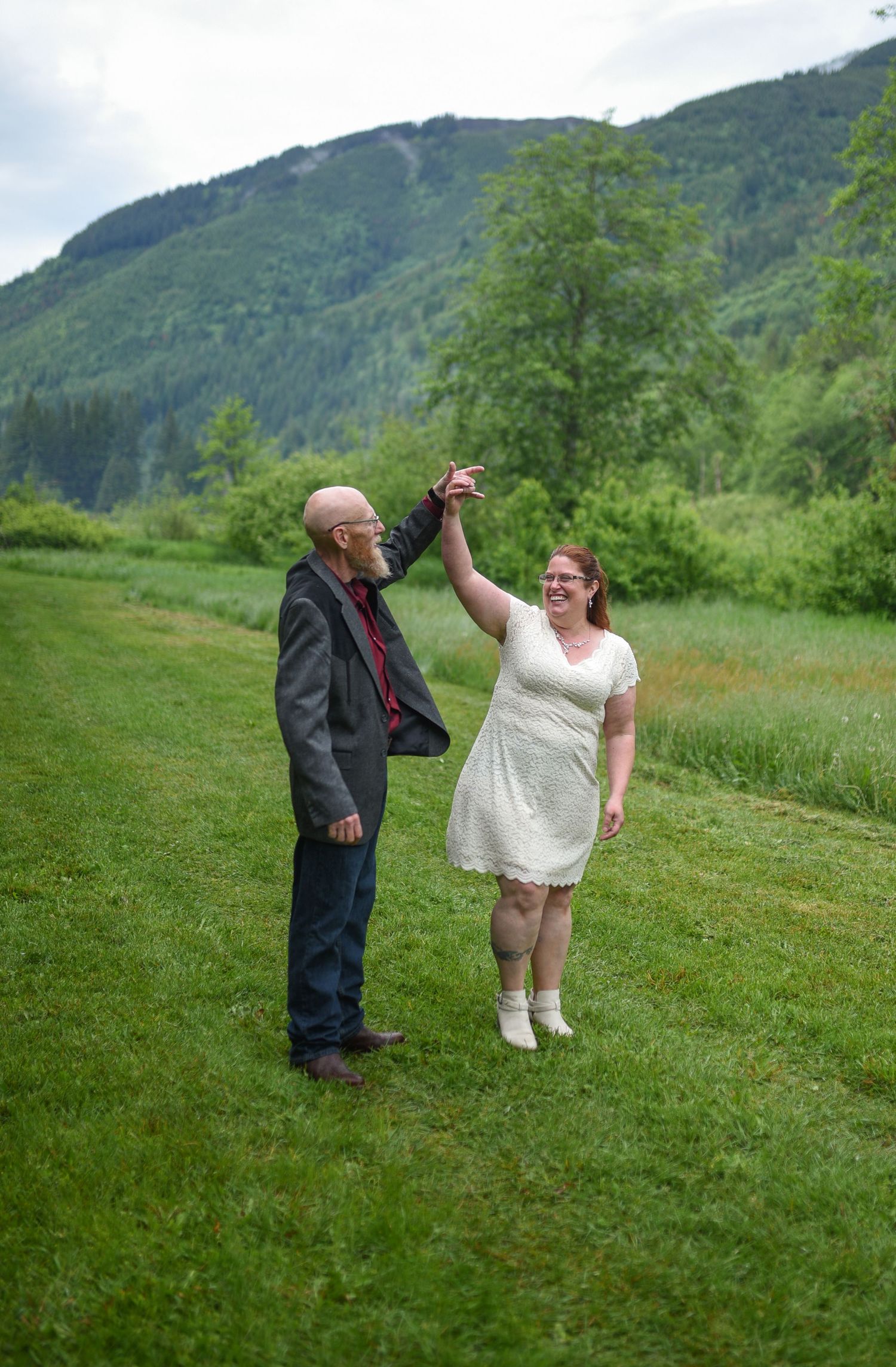 A couple dances outdoors on green grass with lush mountain scenery, the woman in a white dress and man in a dark suit.