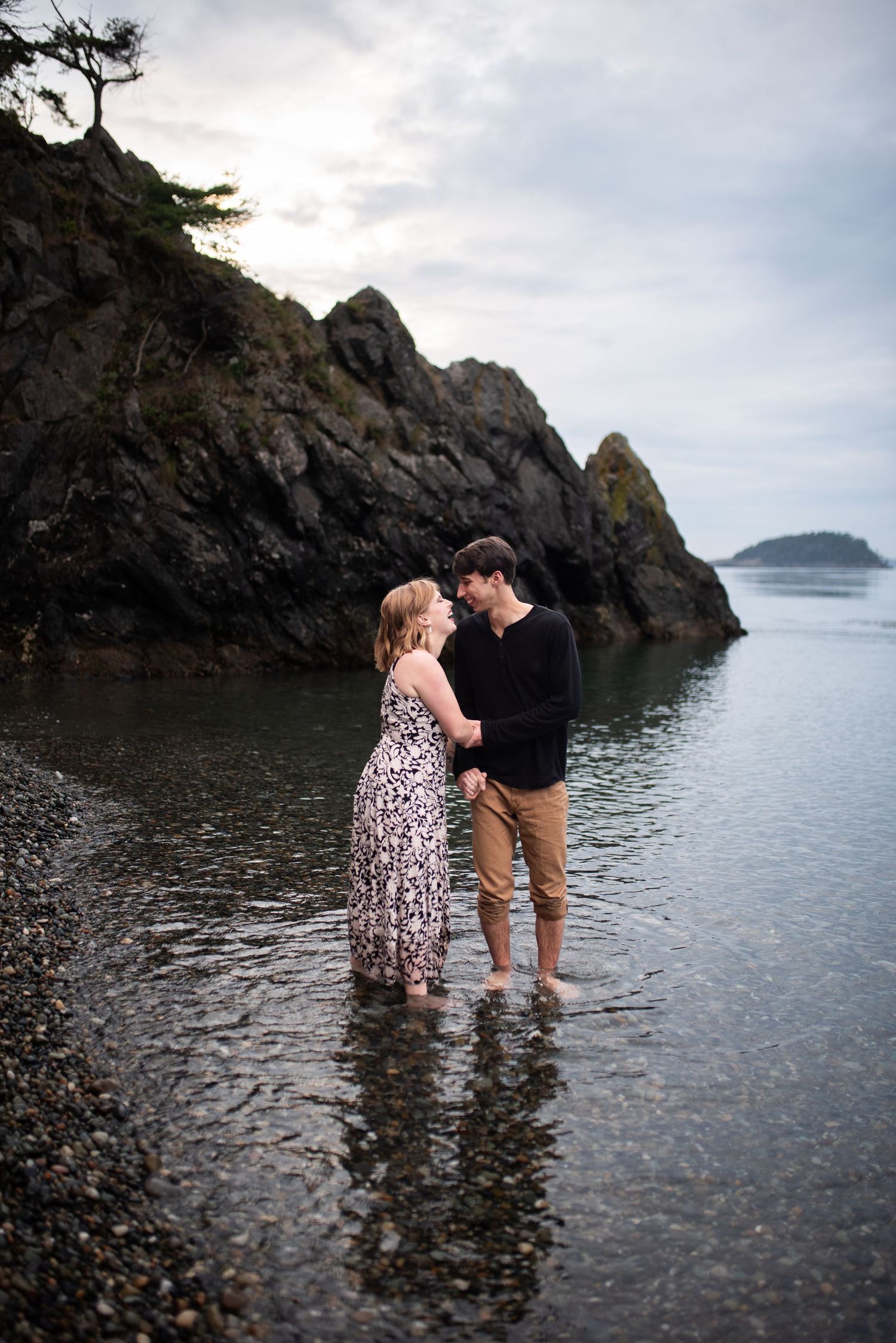 Couple holds hands standing in shallow coastal water with dramatic rocky cliffs behind them.