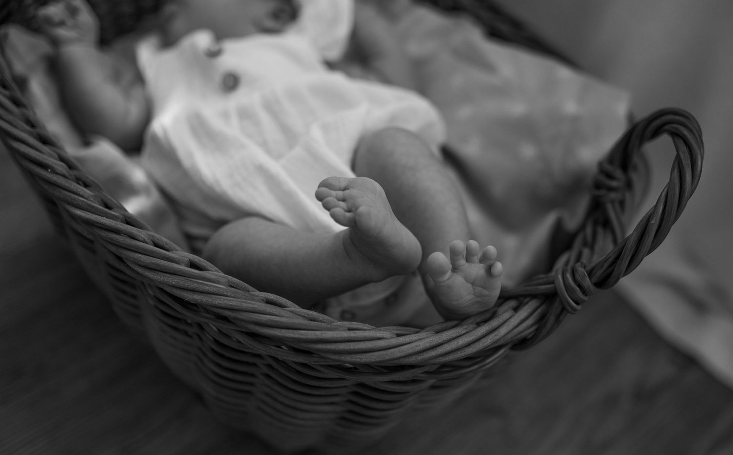 Black and white close-up of a newborn baby lying in a wicker basket wearing a white outfit.