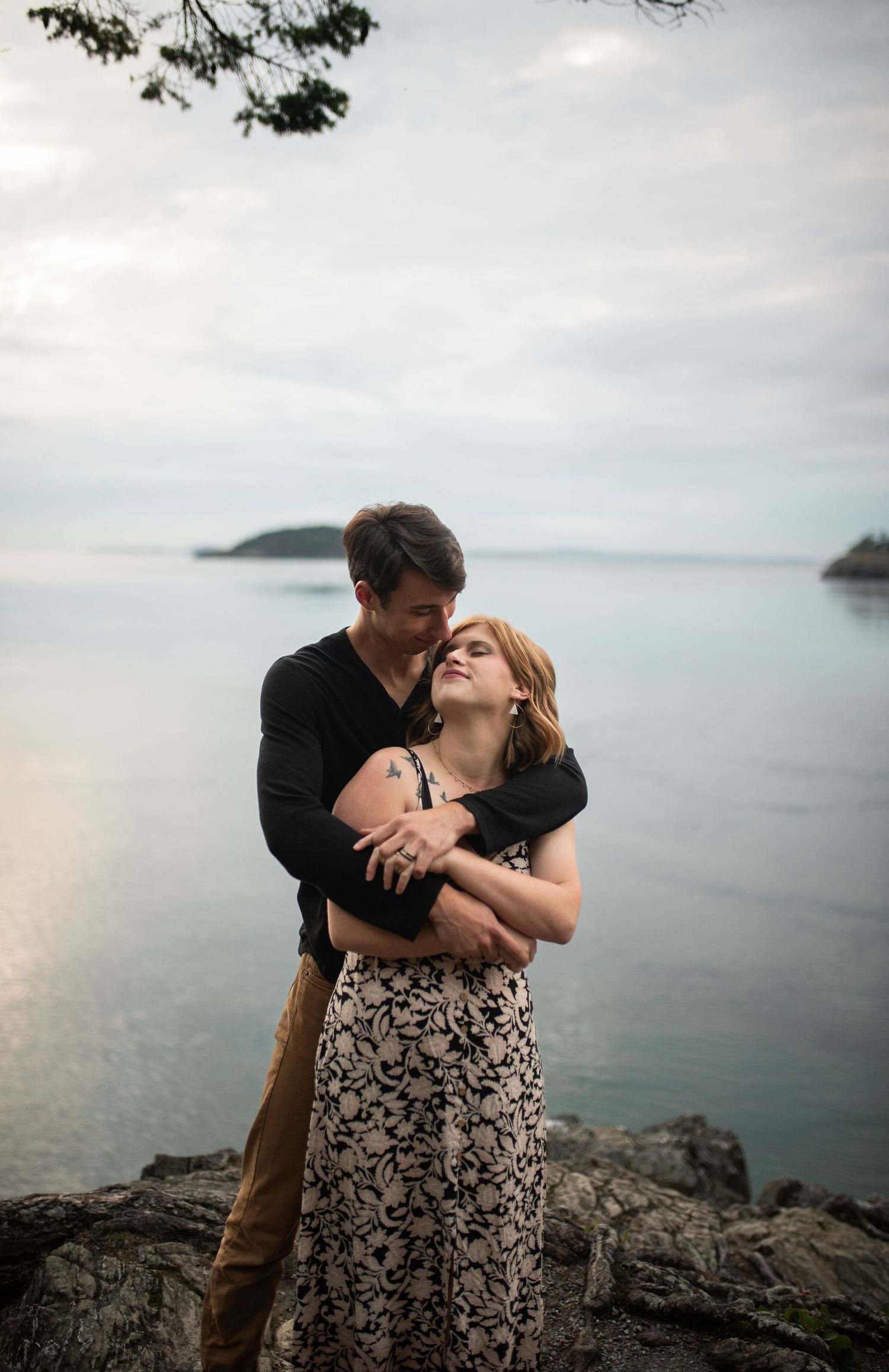 Couple embracing on coastal rocks with calm ocean and small island visible in the soft evening light.