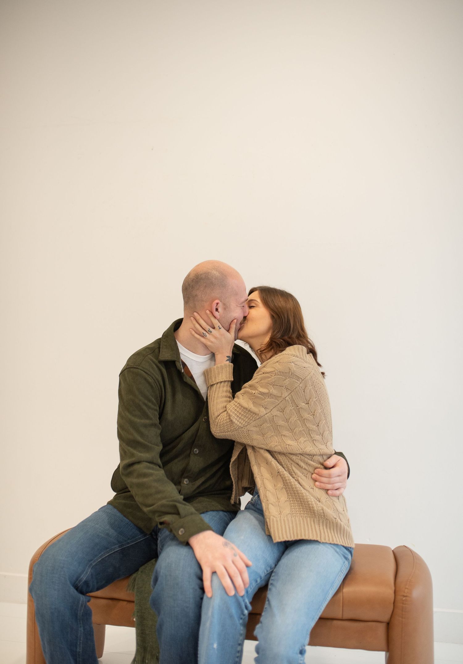 Couple sharing a kiss while seated on a leather bench during an indoor engagement photo session.