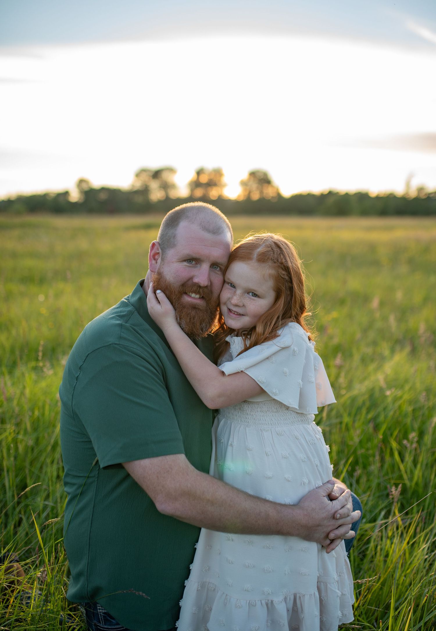 Father with red beard hugs his young red-haired daughter in a green field at golden hour.