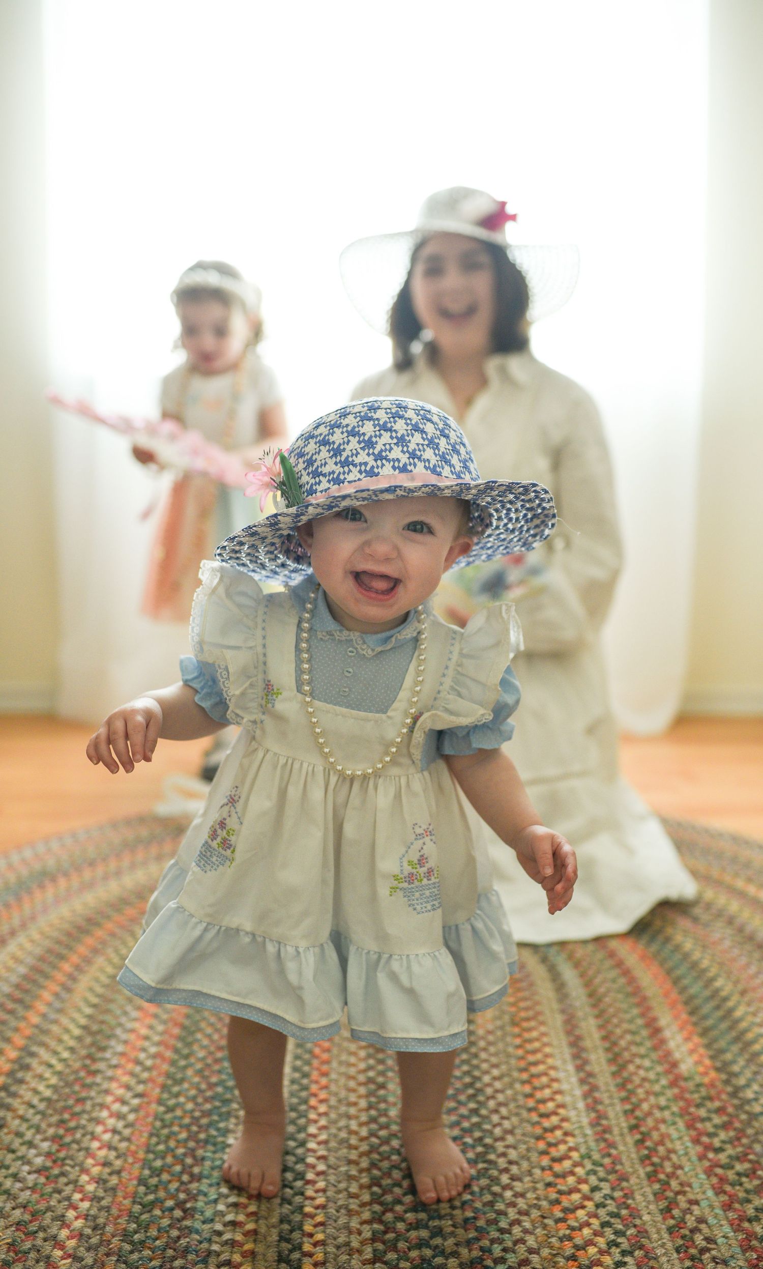An adorable baby in a blue dress and pearl necklace smiles while wearing a houndstooth sun hat.