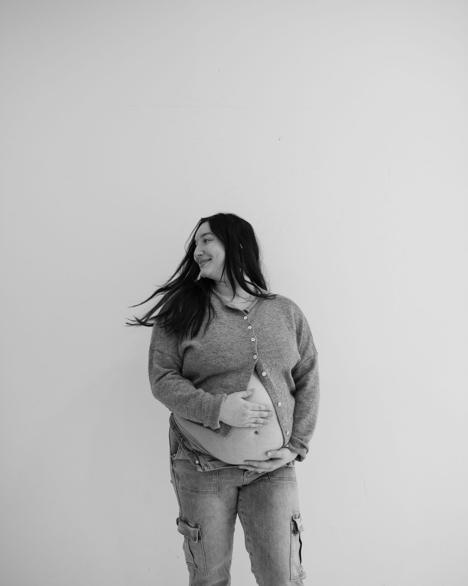 Black and white photo of a smiling pregnant woman cradling her baby bump, wearing jeans and a cardigan.