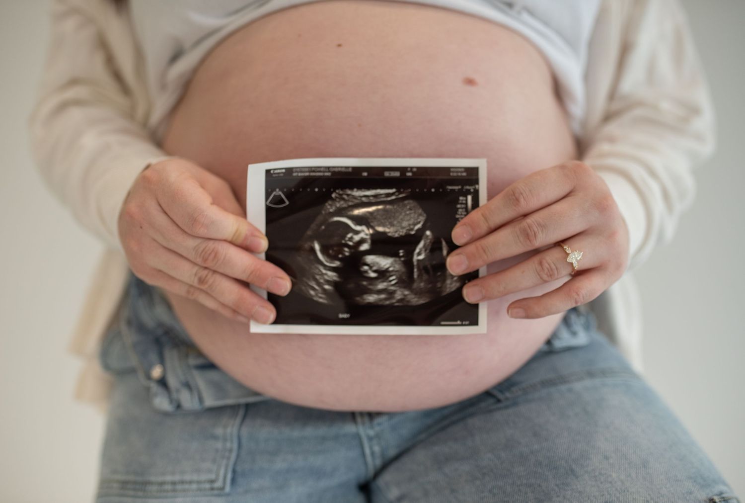 Close-up of pregnant woman holding ultrasound photo in front of her bare baby bump wearing jeans and white cardigan.