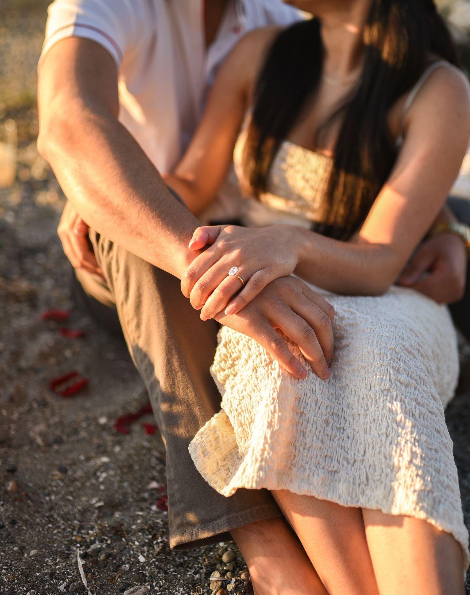 Couple sitting together on a beach, holding hands with an engagement ring visible, wearing casual summer outfits.