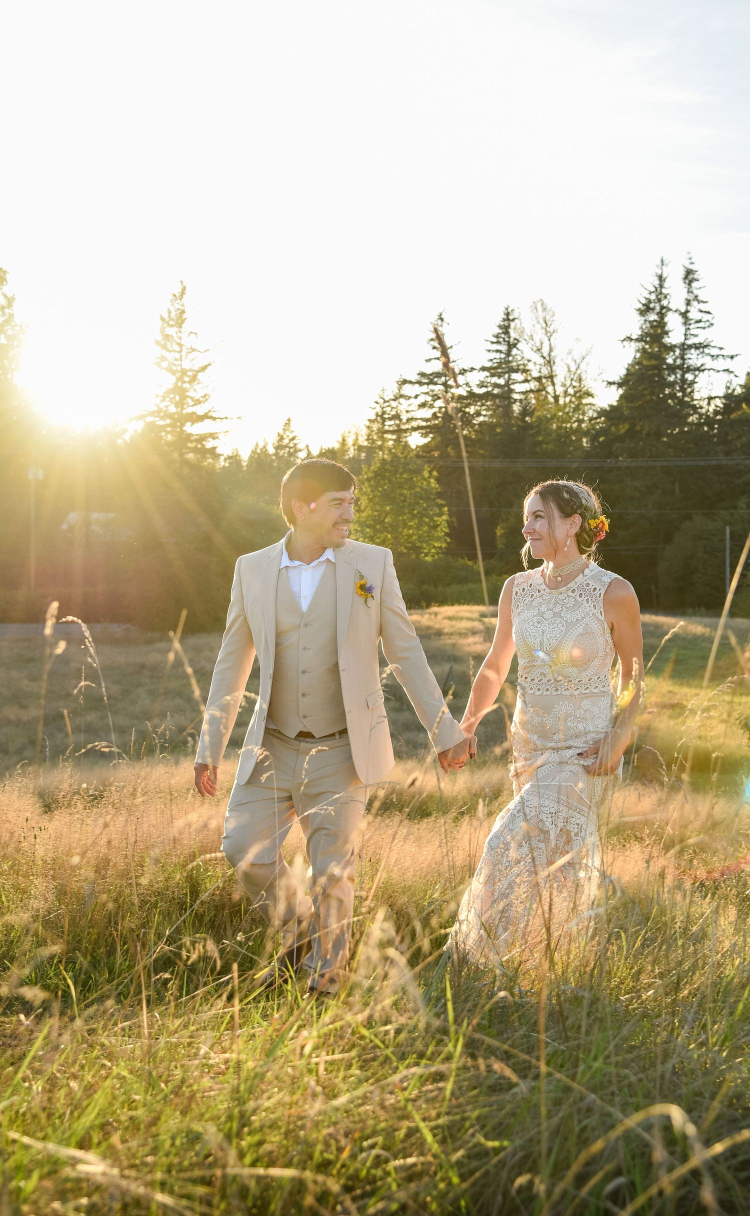Bride and groom holding hands walking through golden field at sunset, wearing elegant beige wedding attire.