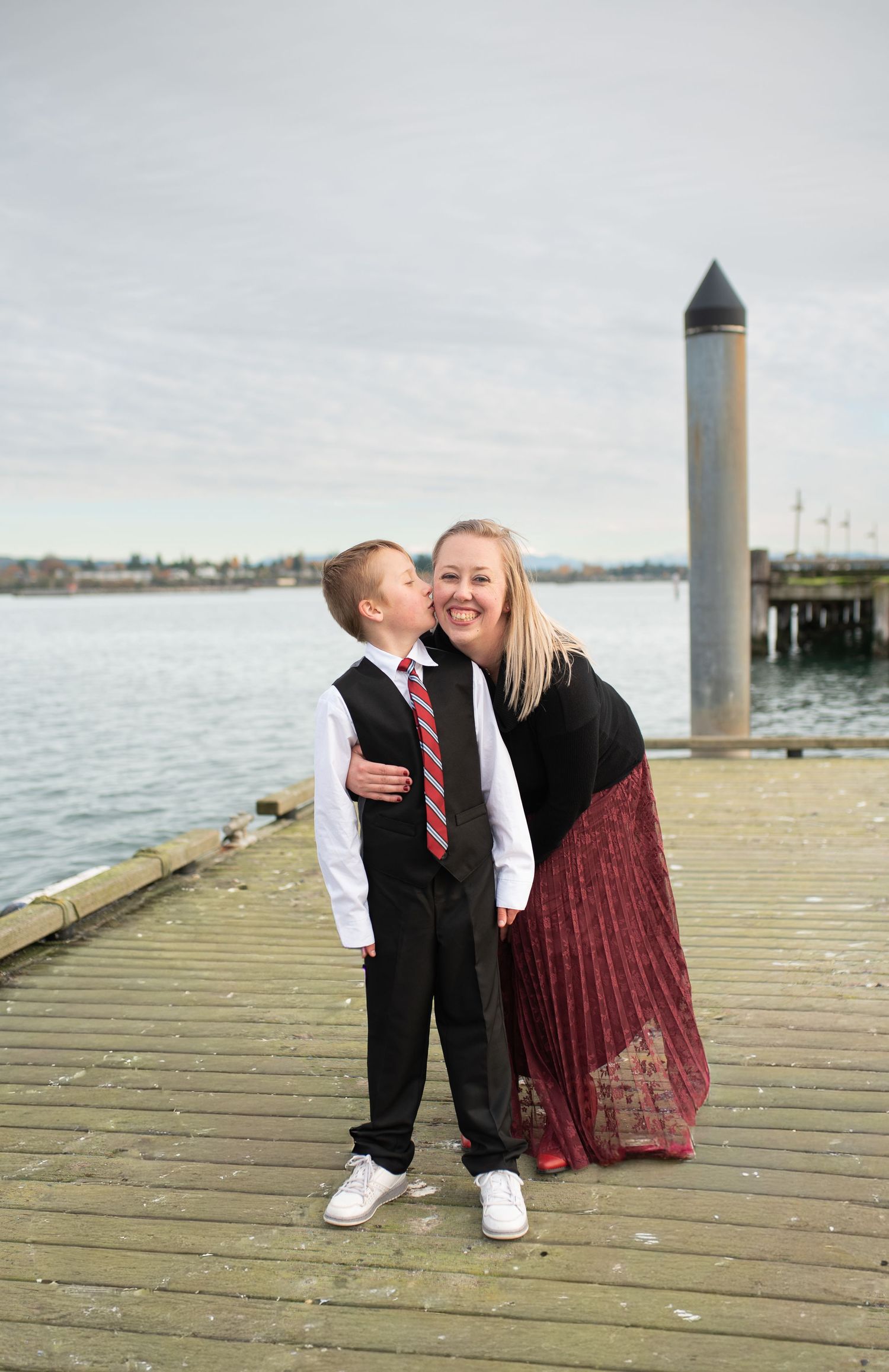 Young boy in suit kissing smiling blonde mother on wooden pier by the water.