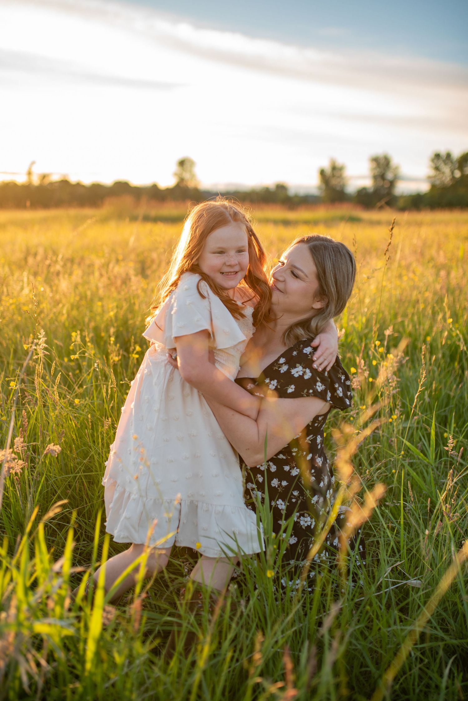 Mother laughs while hugging her red-haired daughter in a bright sunlit green meadow.