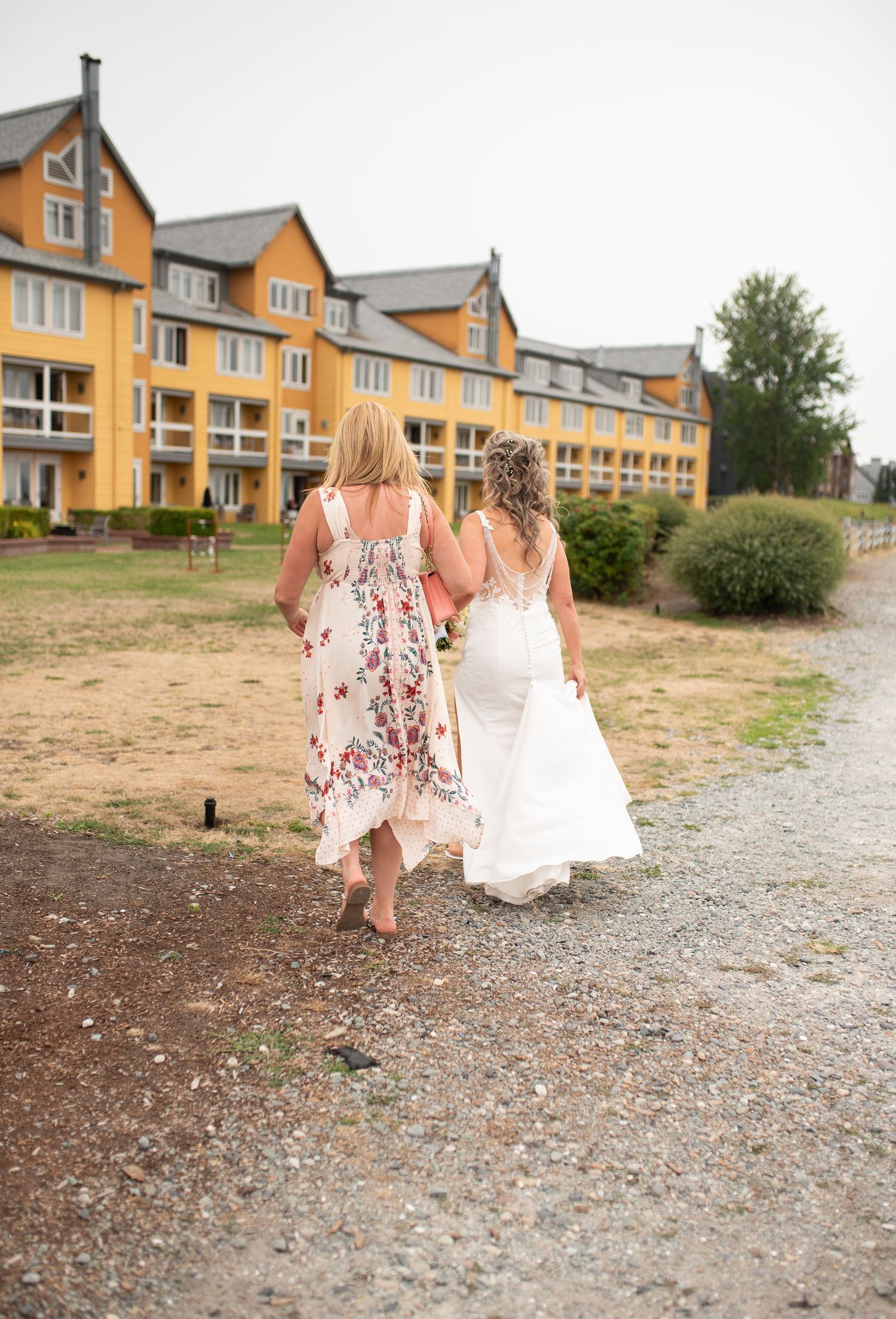 Two women, one in a floral dress and one in a white wedding gown, walk together toward a yellow hotel building.