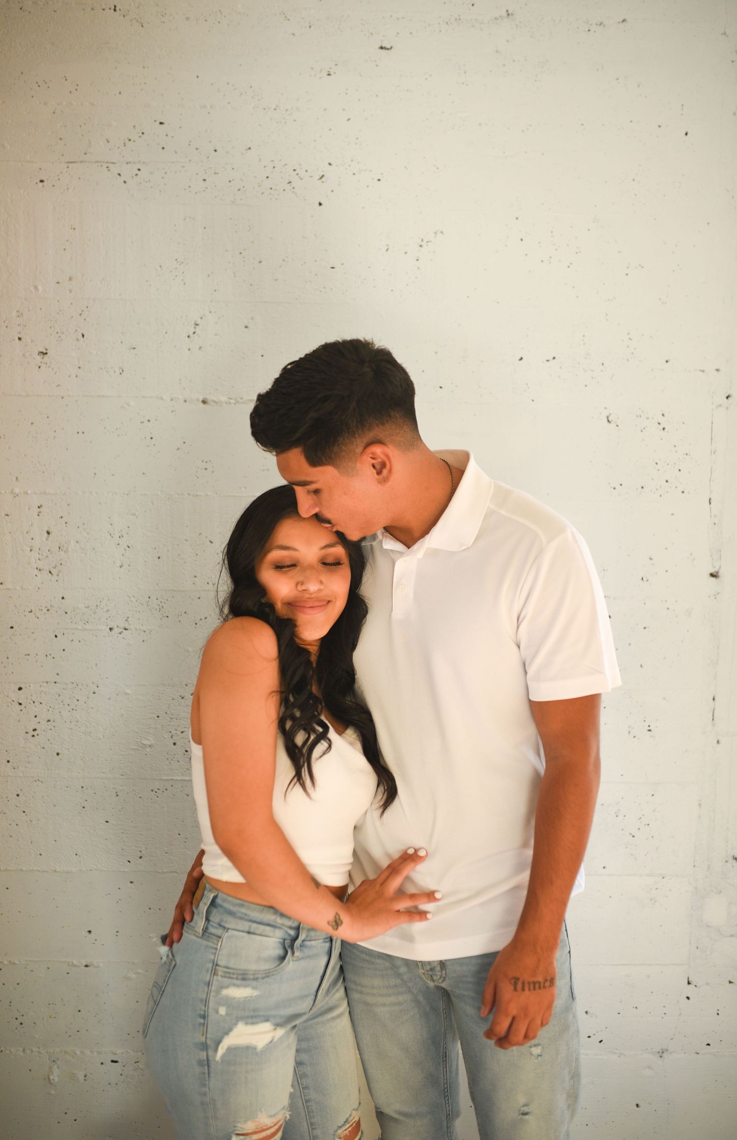 Couple in matching white tops and jeans sharing a tender moment against a light concrete wall.