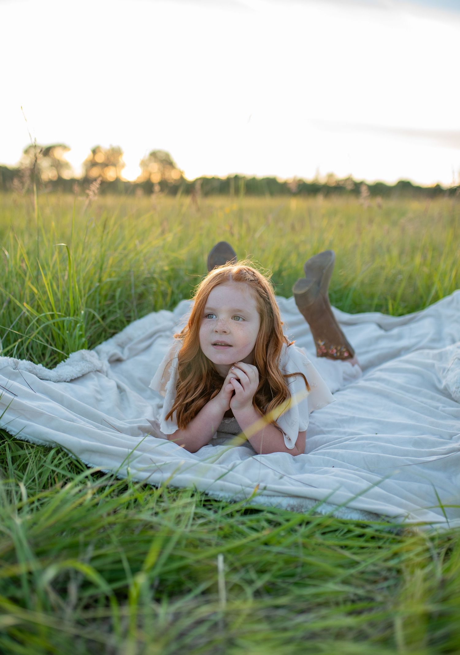 Young red-haired girl in white dress lies on a blanket in a green field at golden hour.