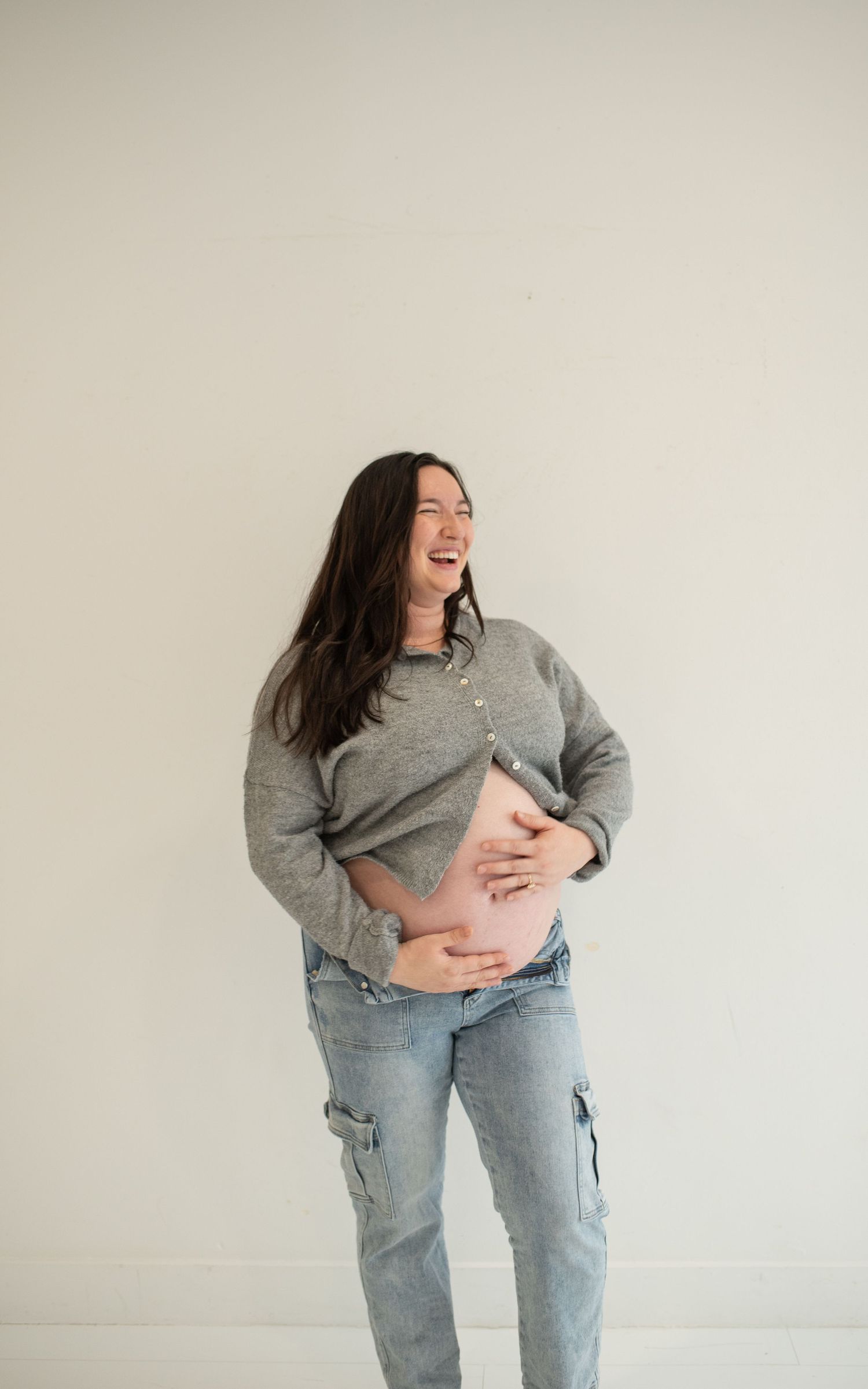 Laughing pregnant woman in gray sweater and jeans cradling baby bump against white wall.