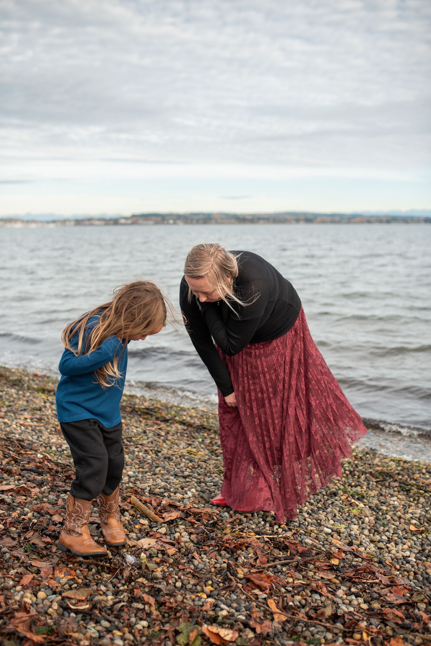 Mother bending down to interact with young child on rocky pebble beach shoreline.