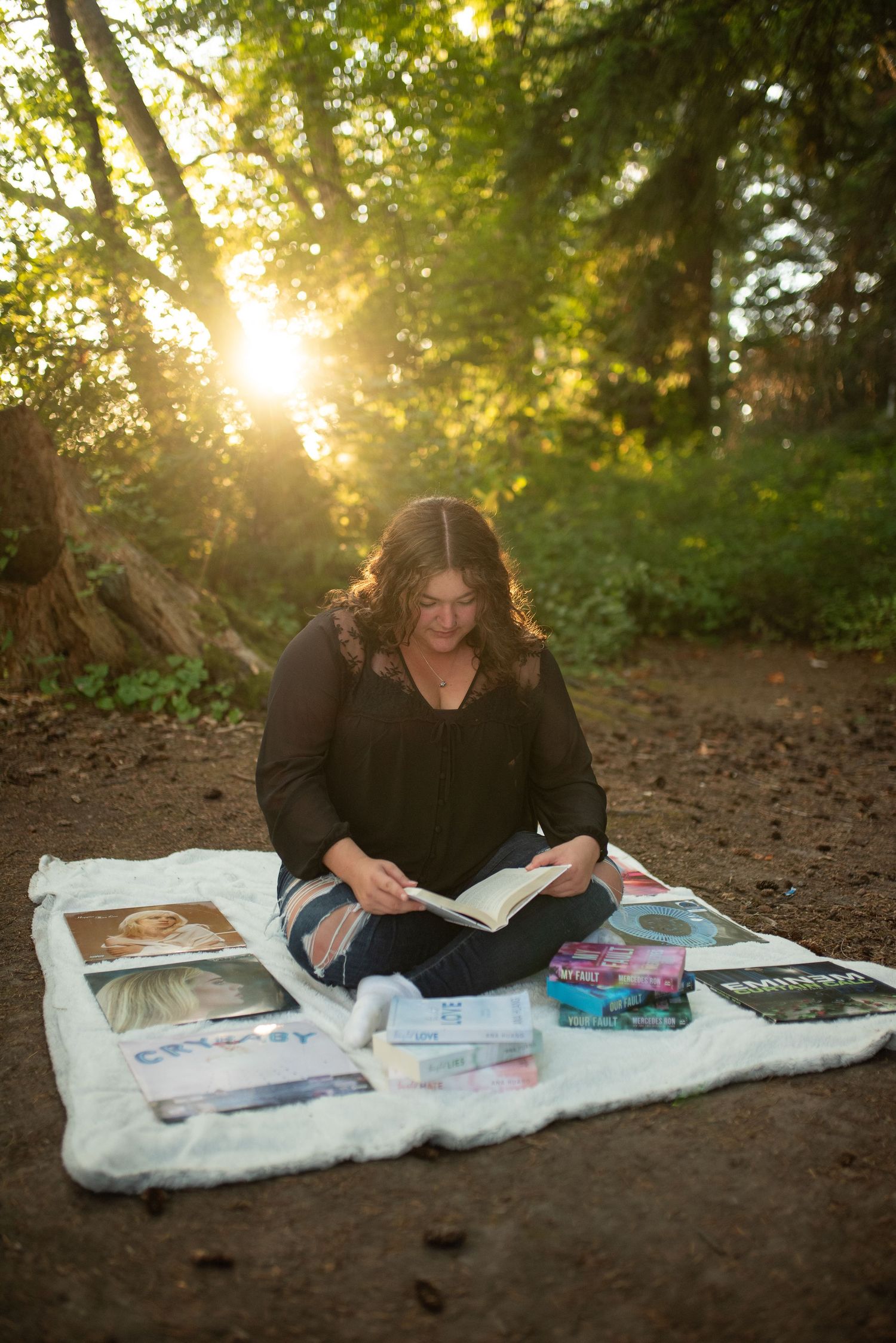 Teen girl sits cross-legged reading surrounded by books in sunlit forest.